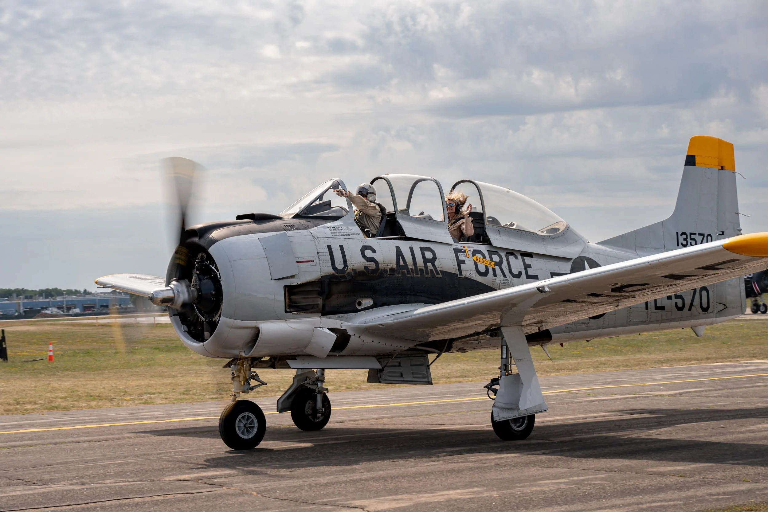 A vintage U.S. Air Force aircraft with a single jet engine, parked on a runway, with two people in the cockpit. One person is wearing a flight suit and helmet, pointing forward, and the other is wearing sunglasses, sitting in the passenger seat. The 