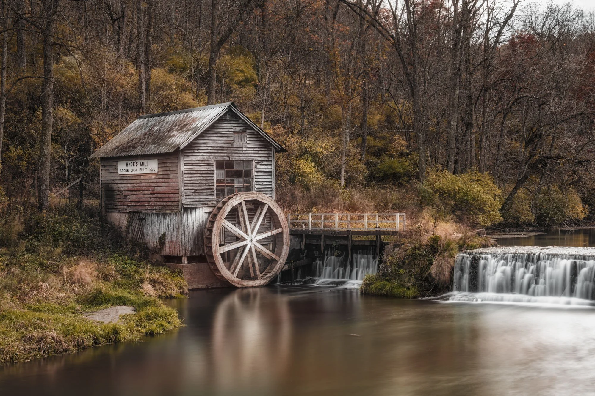 An old wooden watermill with a large water wheel beside a river, surrounded by fall foliage and leafless trees.