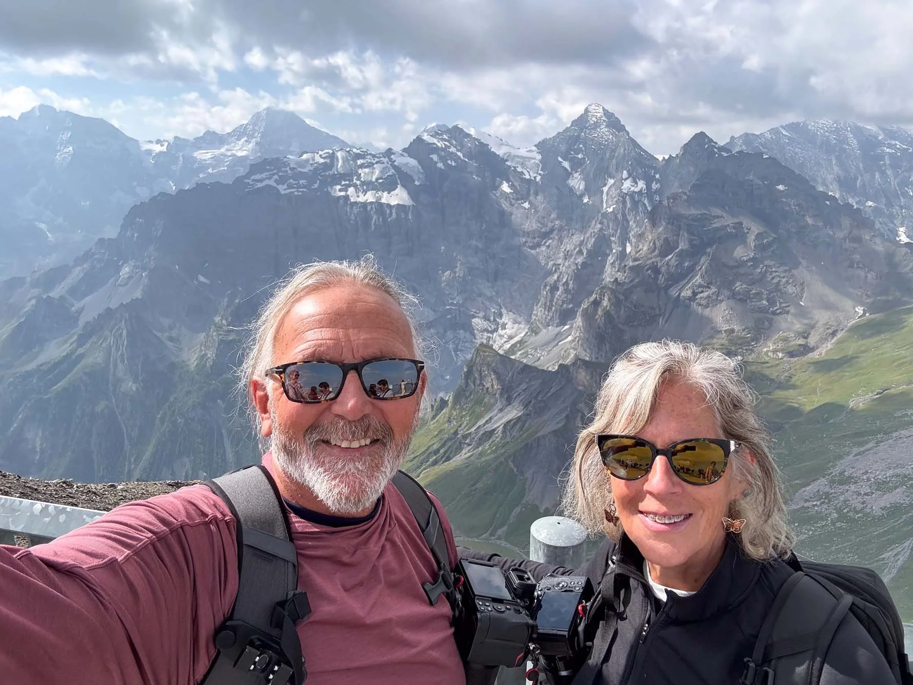 A smiling older man and woman taking a selfie in front of a mountainous landscape with snow-capped peaks, green slopes, and a partly cloudy sky.