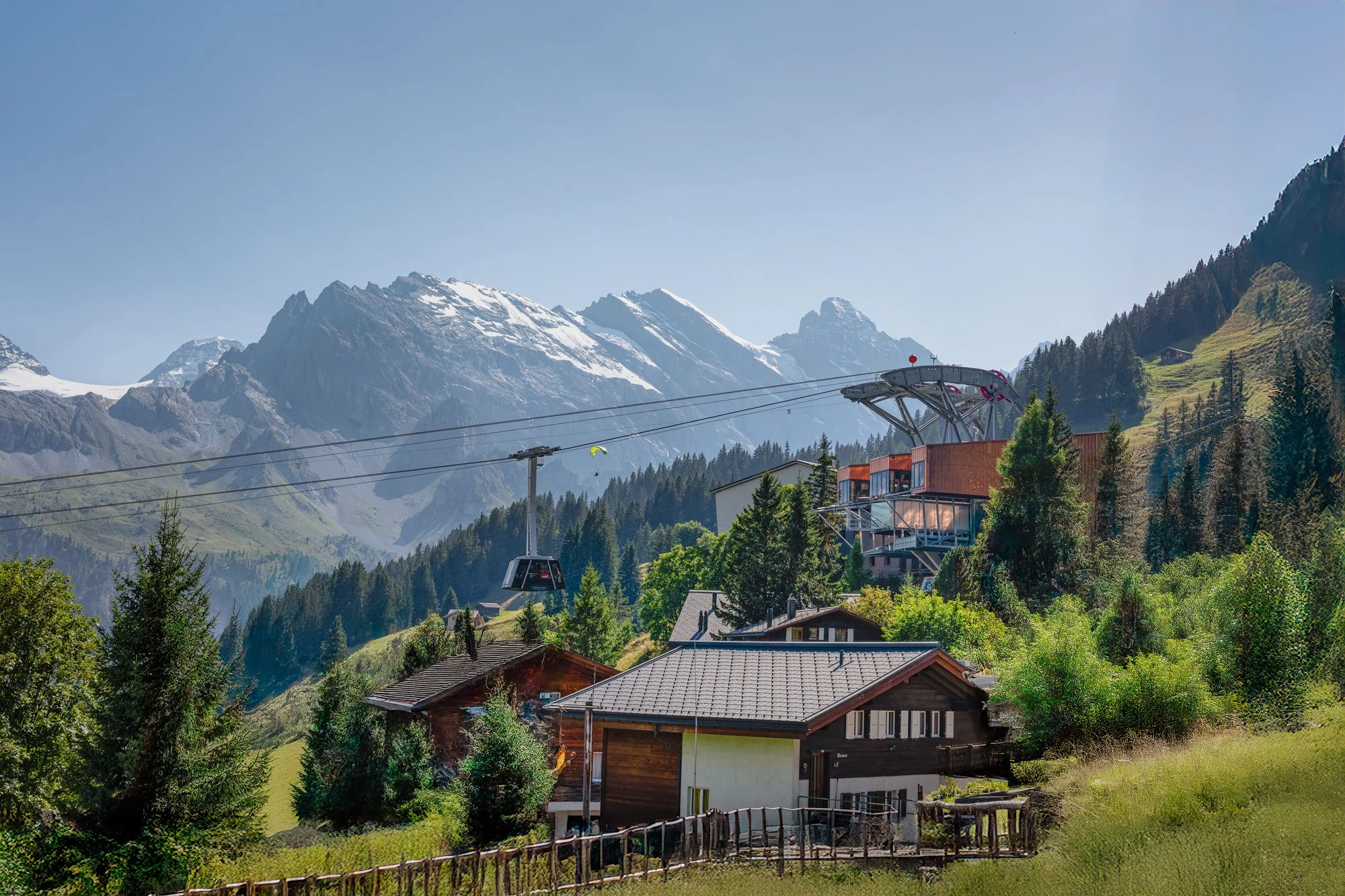 Mountain landscape with chalet houses, trees, and a ski lift in a lush green valley with snow-capped peaks in the background.