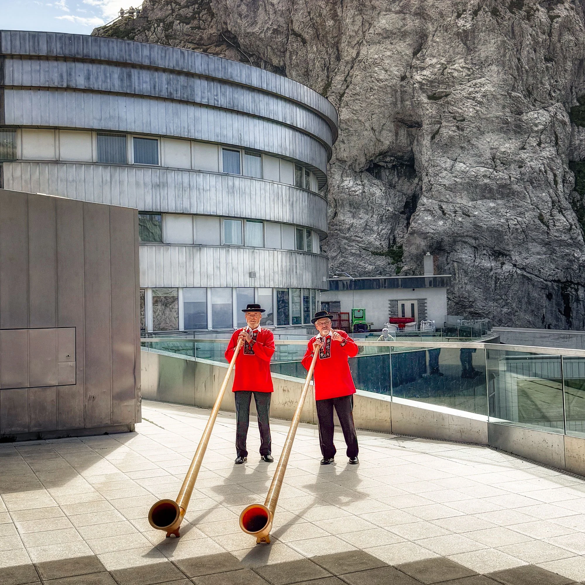 Two men in traditional red costumes and black hats are playing alphorns on a tiled outdoor terrace with glass railings. Behind them is a modern round building against a rocky mountain background.