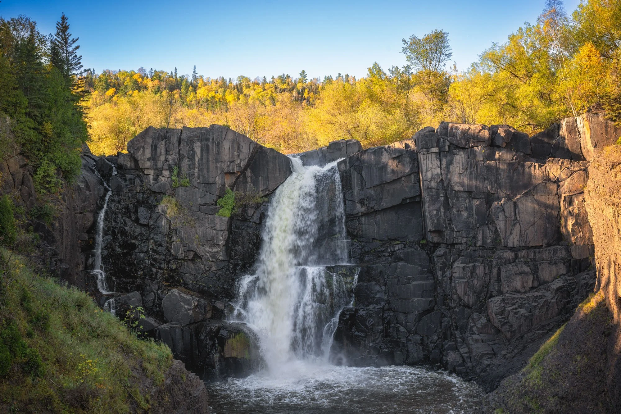 A waterfall flowing over black rocks surrounded by trees with fall foliage and a clear blue sky.