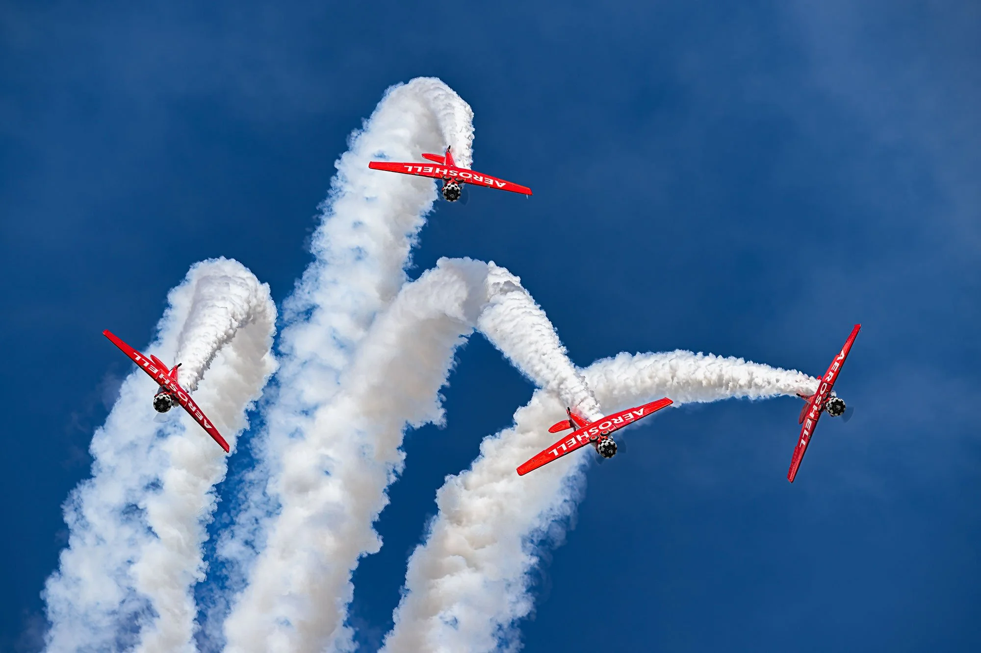 Four red aerobatic planes with 'AEROSHELL' written on them perform an inverted flying stunt, leaving trails of white smoke against a blue sky.