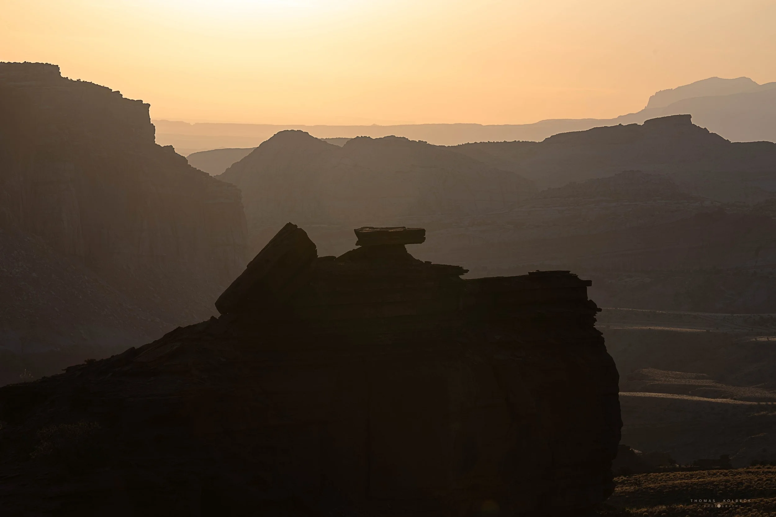 A silhouette of rock formations and mesas during sunset or sunrise in a desert landscape with layered mountains in the background.