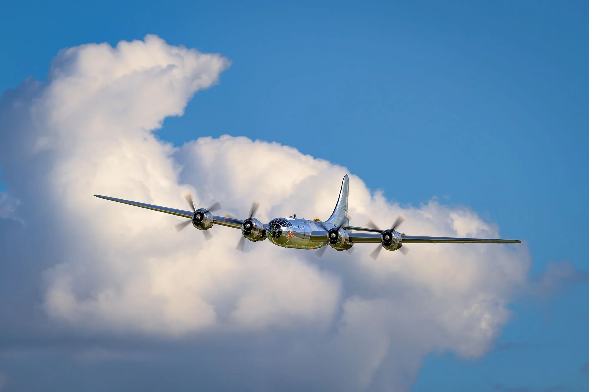 A vintage silver propeller airplane flying in a clear blue sky with large white clouds.