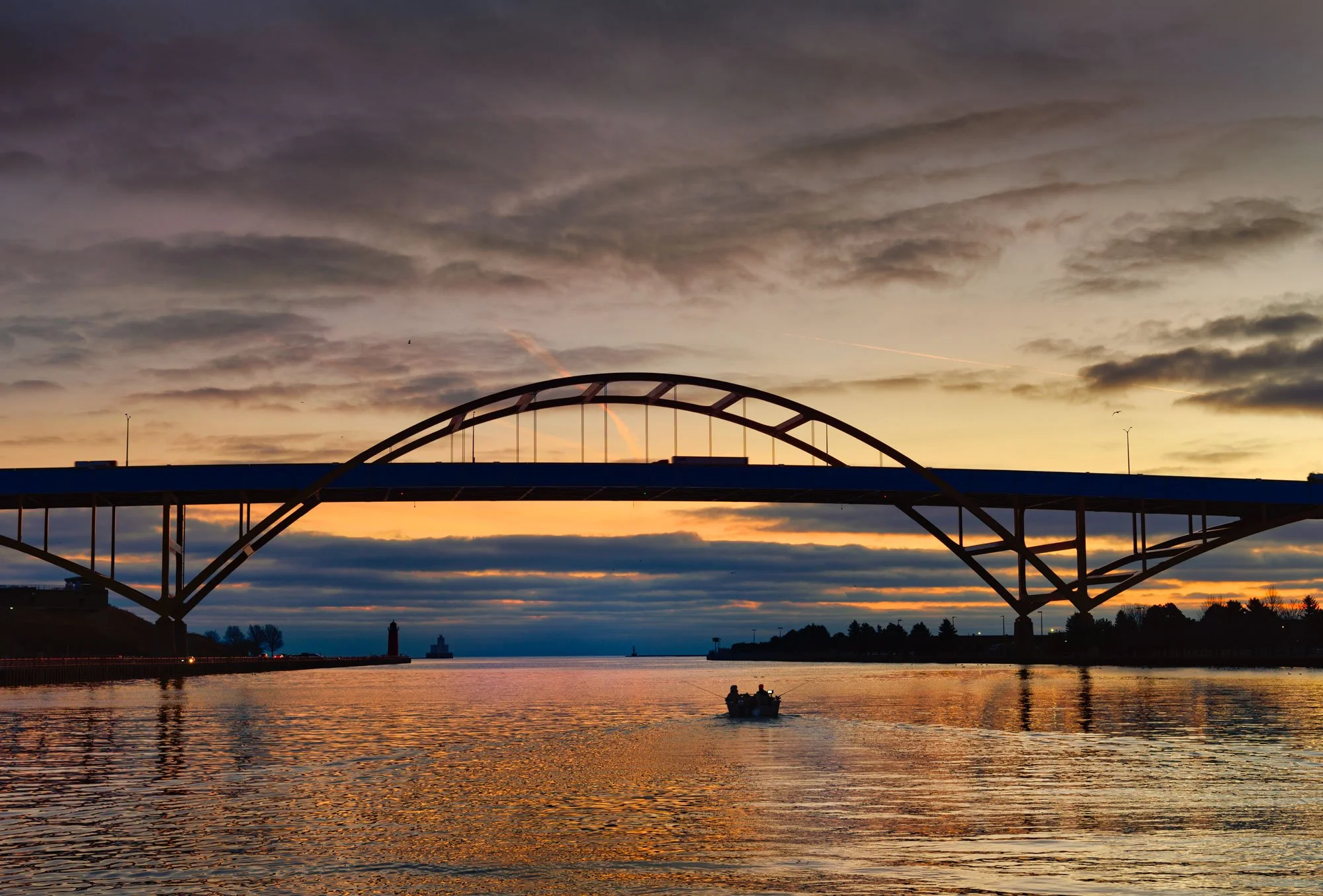 A bridge over a body of water at sunset with a small boat in the water and a cityscape in the background.