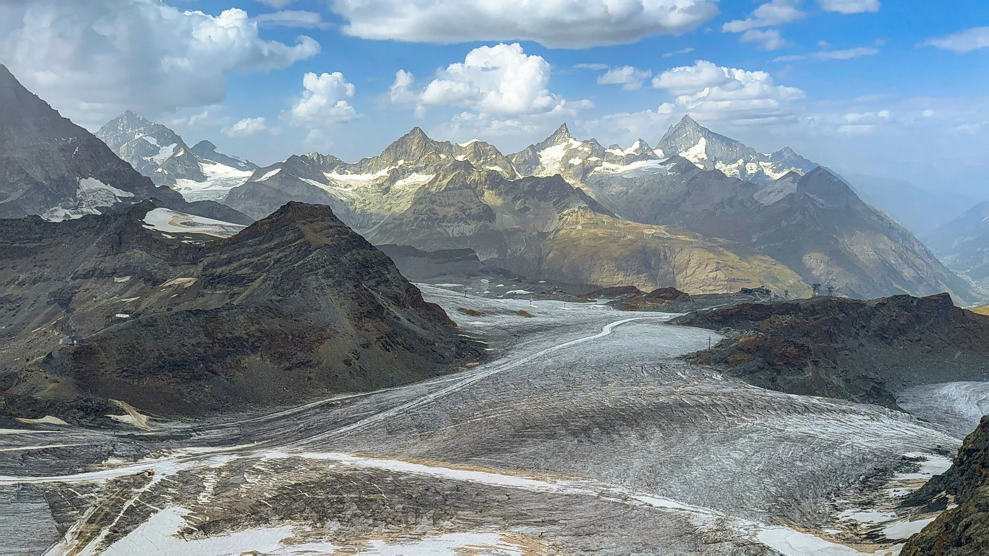 Mountain landscape with snow-capped peaks, rugged terrain, and cloudy sky.