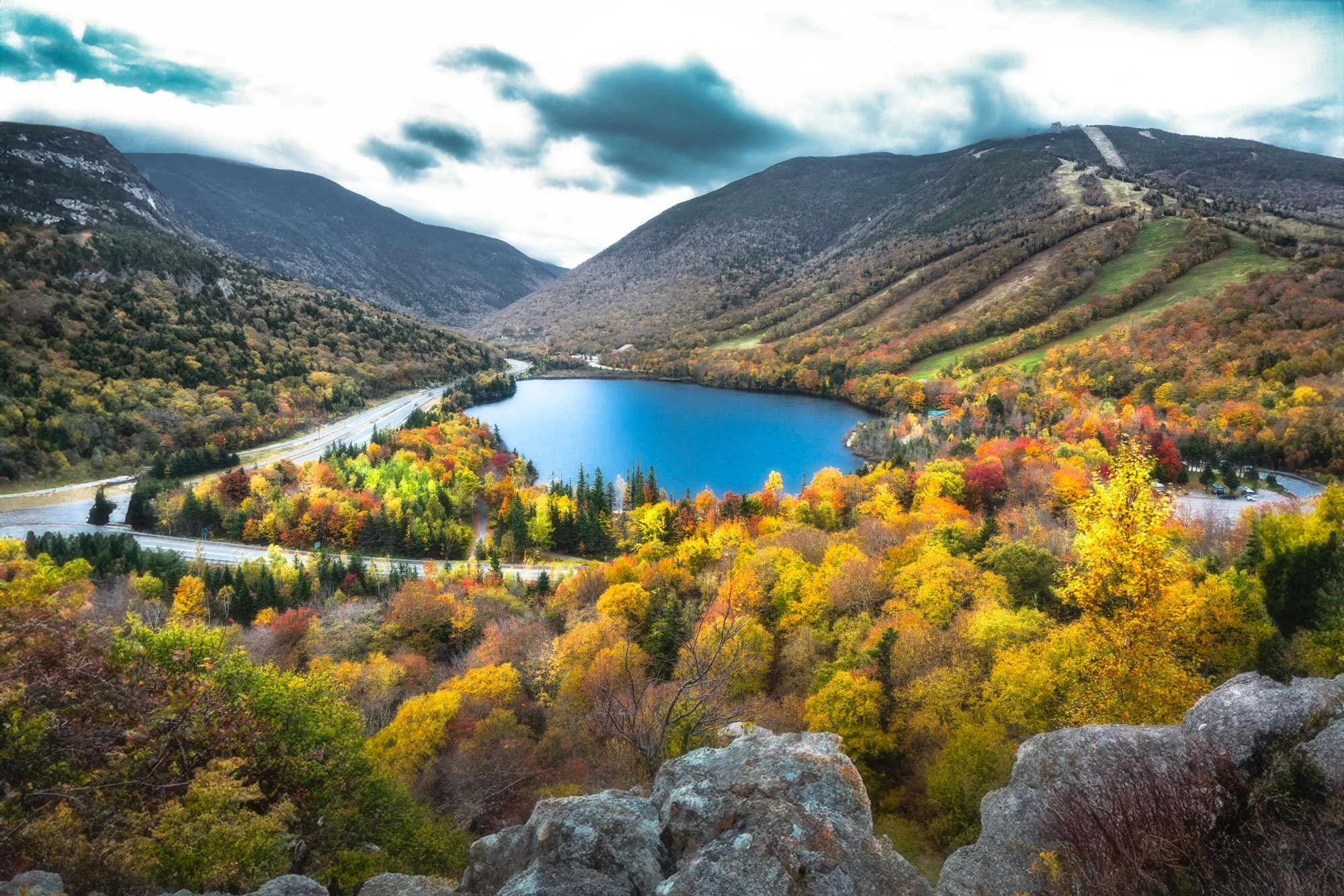 Scenic view of a mountain valley with a blue lake, surrounded by colorful autumn trees, rocky foreground, hills, and mountains under a cloudy sky.