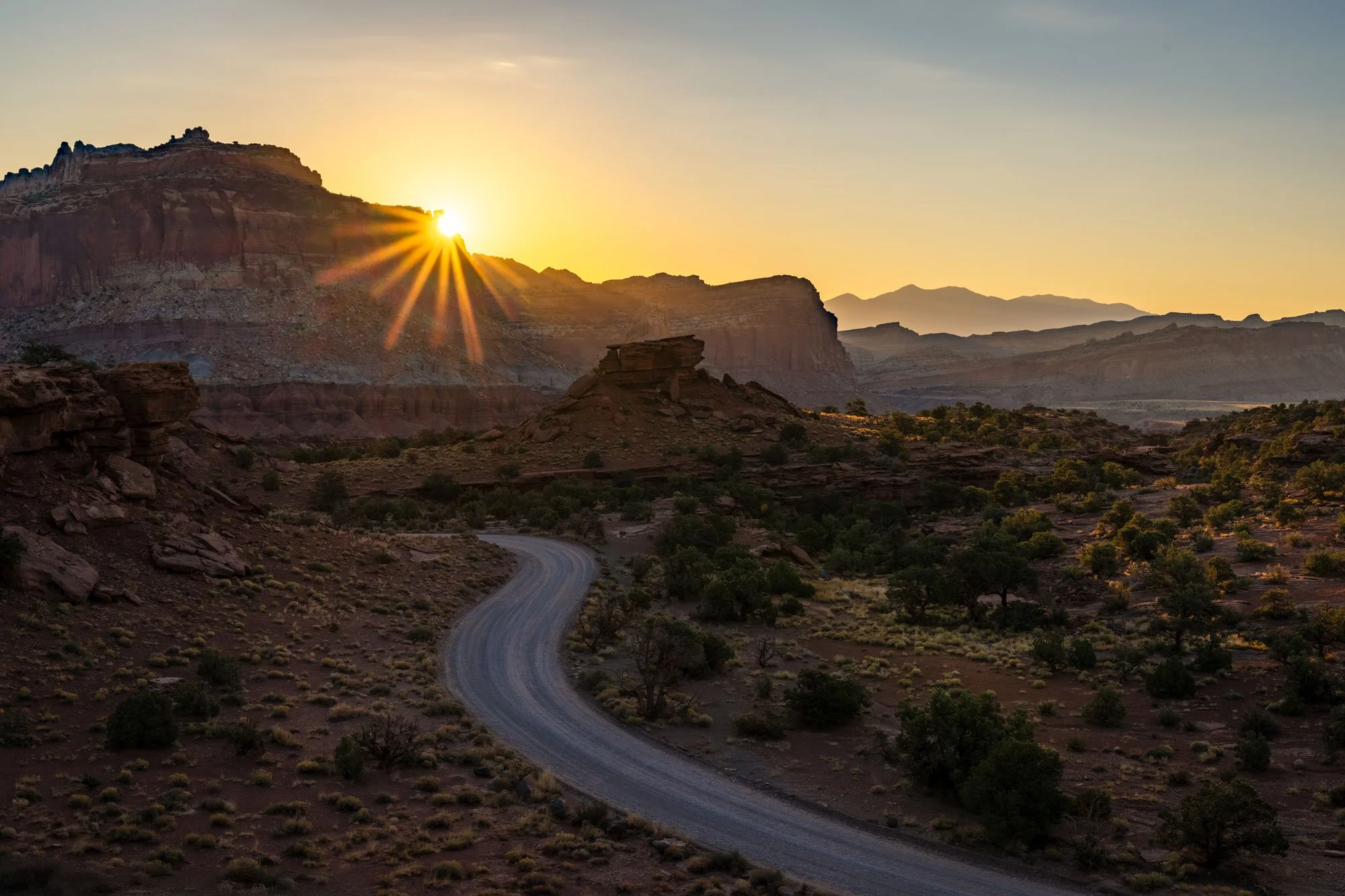 Sunset over a desert canyon with a winding dirt road, rock formations, and distant mountains.