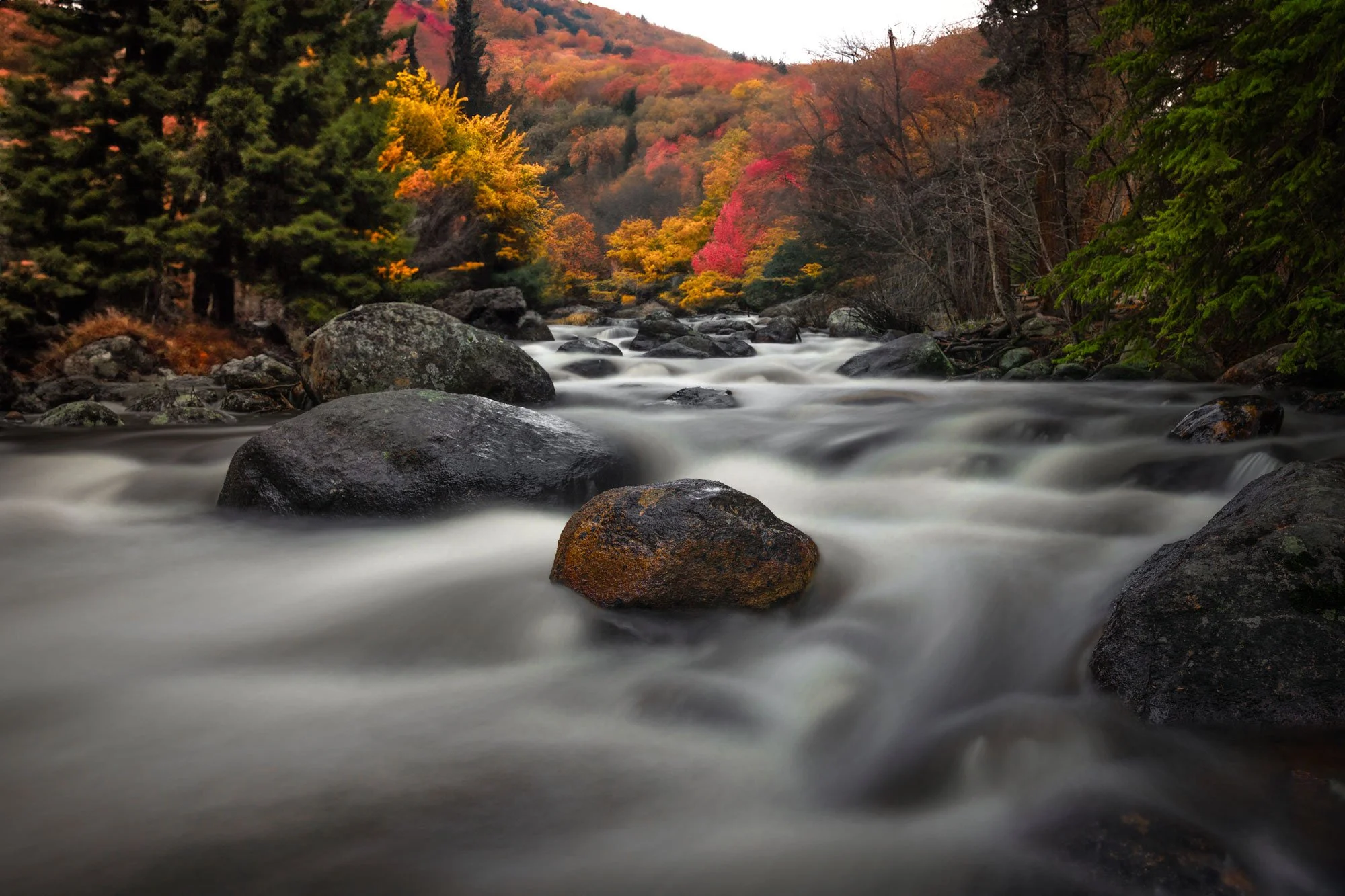 A flowing river with large rocks, surrounded by colorful autumn trees and hills in the background.