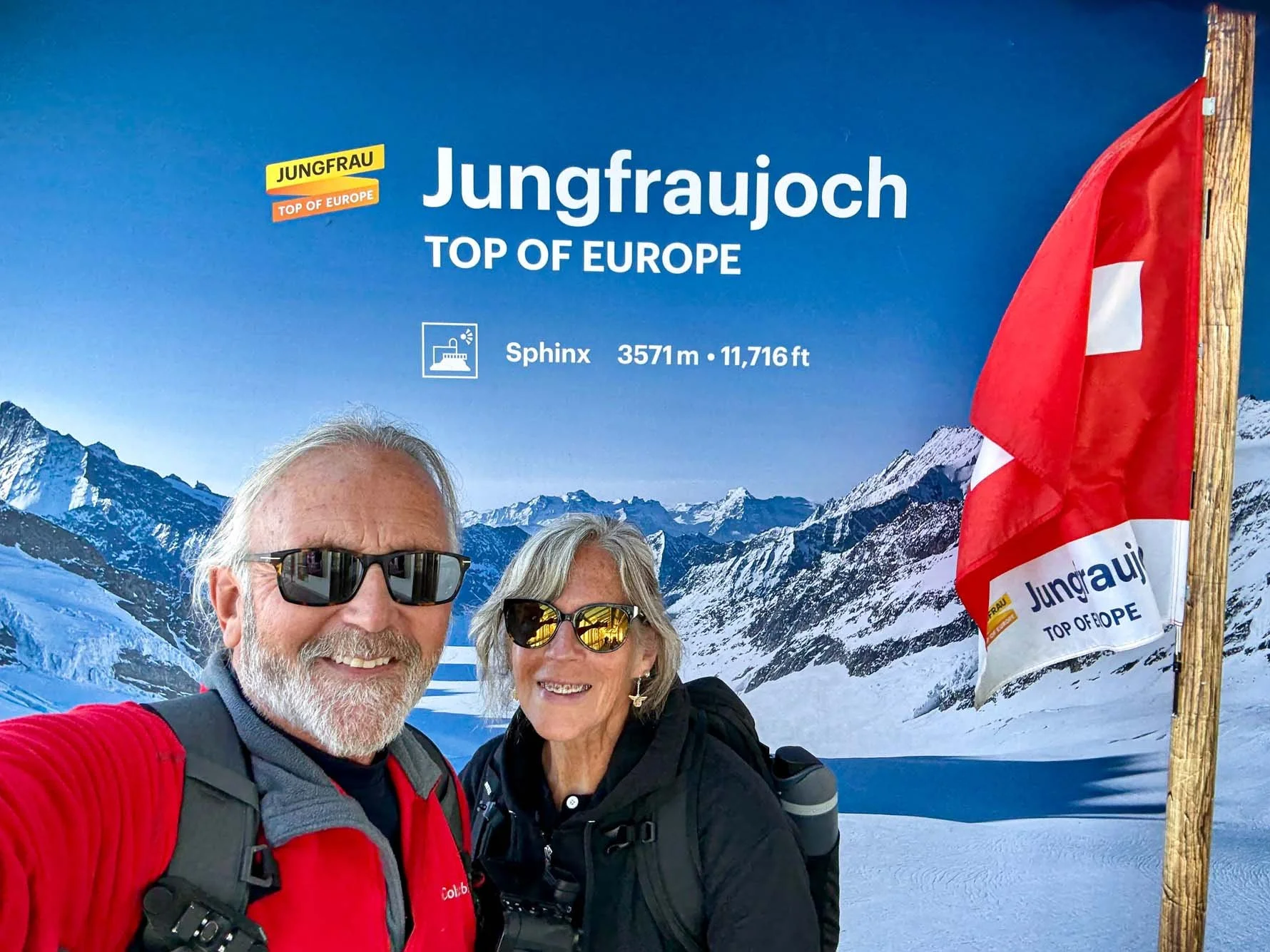 Two smiling hikers wearing sunglasses taking a selfie in front of a sign for Jungfraujoch, the top of Europe, at an altitude of 11,716 feet, with snowy mountains in the background and a Swiss flag next to the sign.
