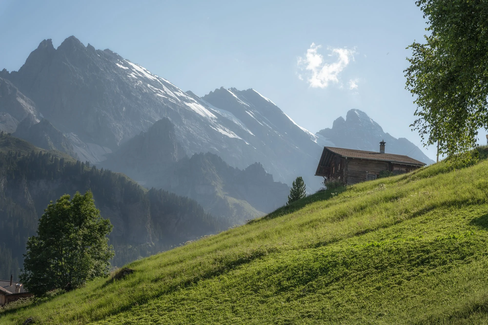 A wooden cabin on a grassy hillside with large snow-capped mountains in the background and a partly cloudy sky.