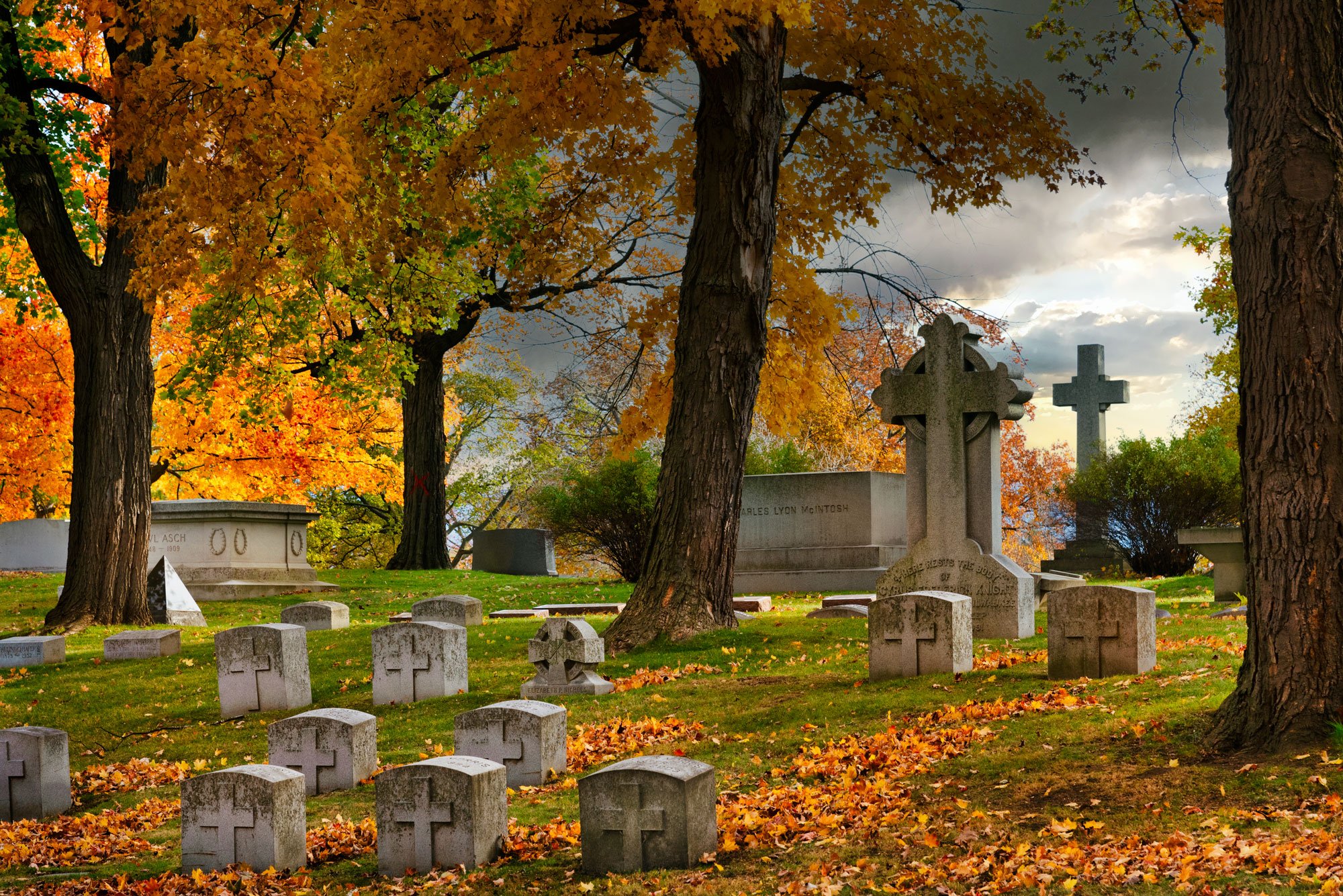 A graveyard with various tombstones and crosses among trees with vibrant fall foliage, under a cloudy sky.