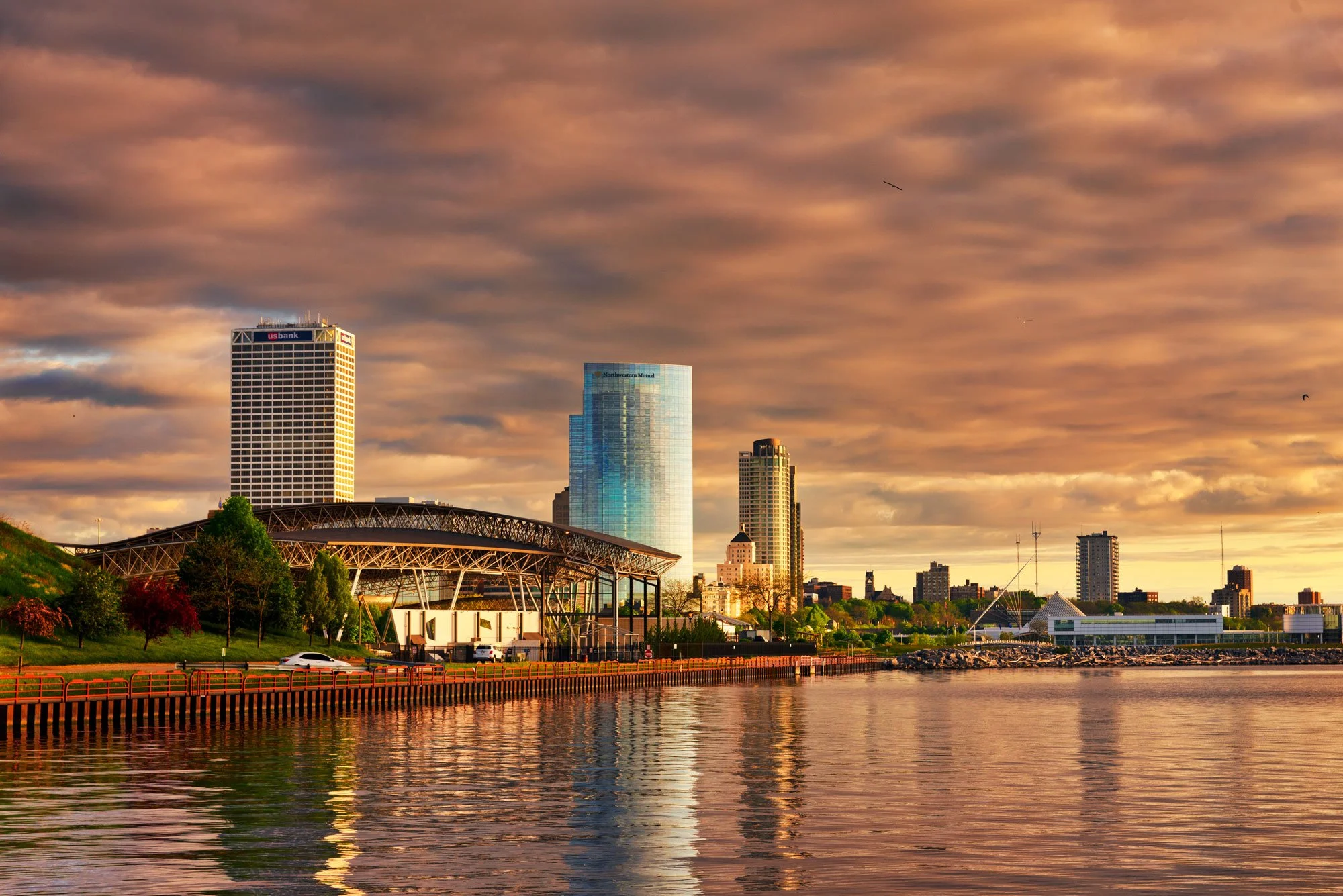 City skyline with tall buildings near a waterfront during sunset, with colorful clouds overhead and reflections in the water.