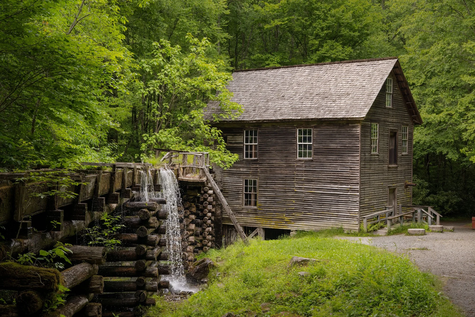 A rustic wooden watermill surrounded by lush green trees with water flowing over the mill's wooden wheel.