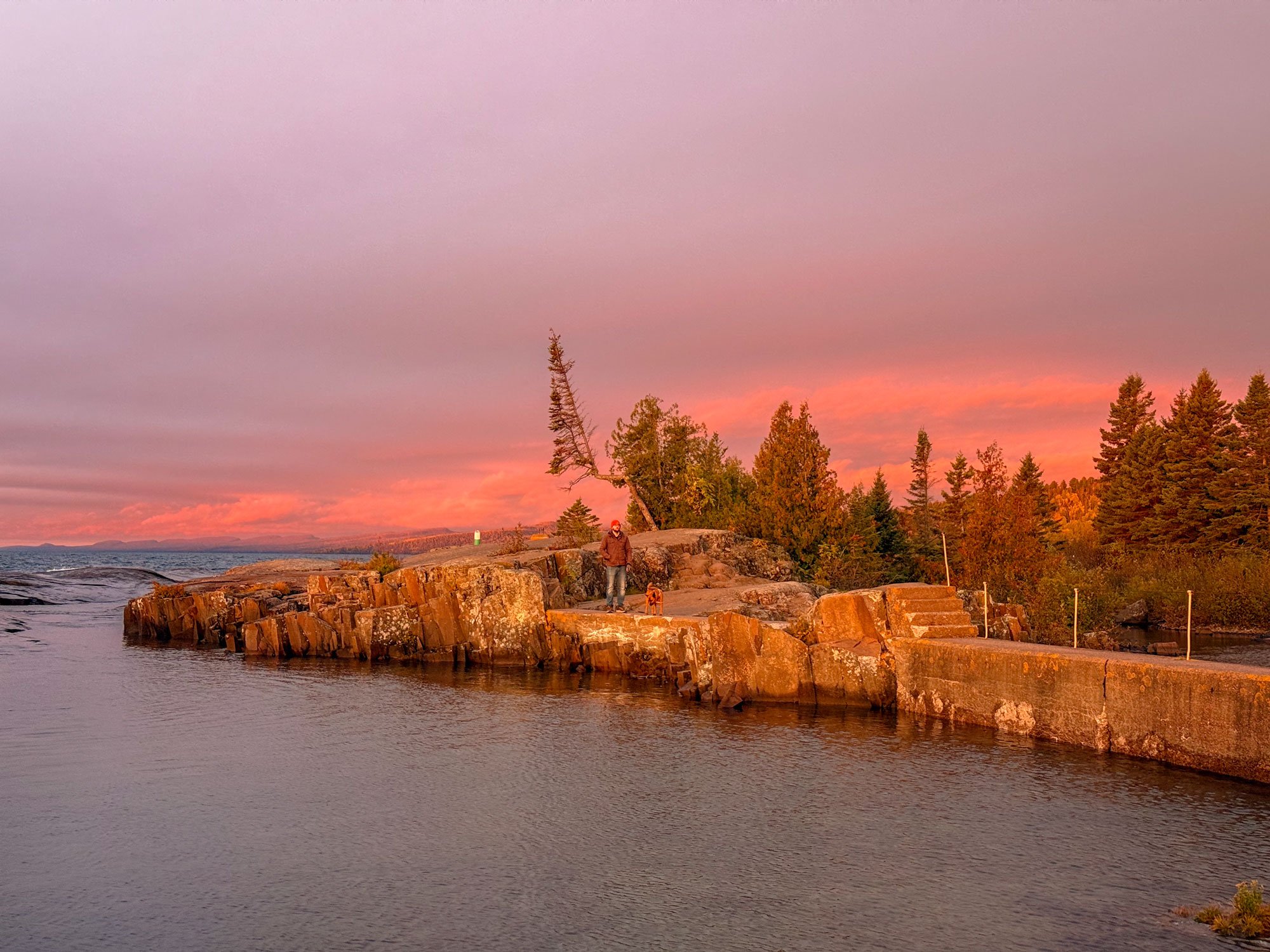 A man and dog standing on a rocky shoreline during sunset, with colorful sky and trees in the background.