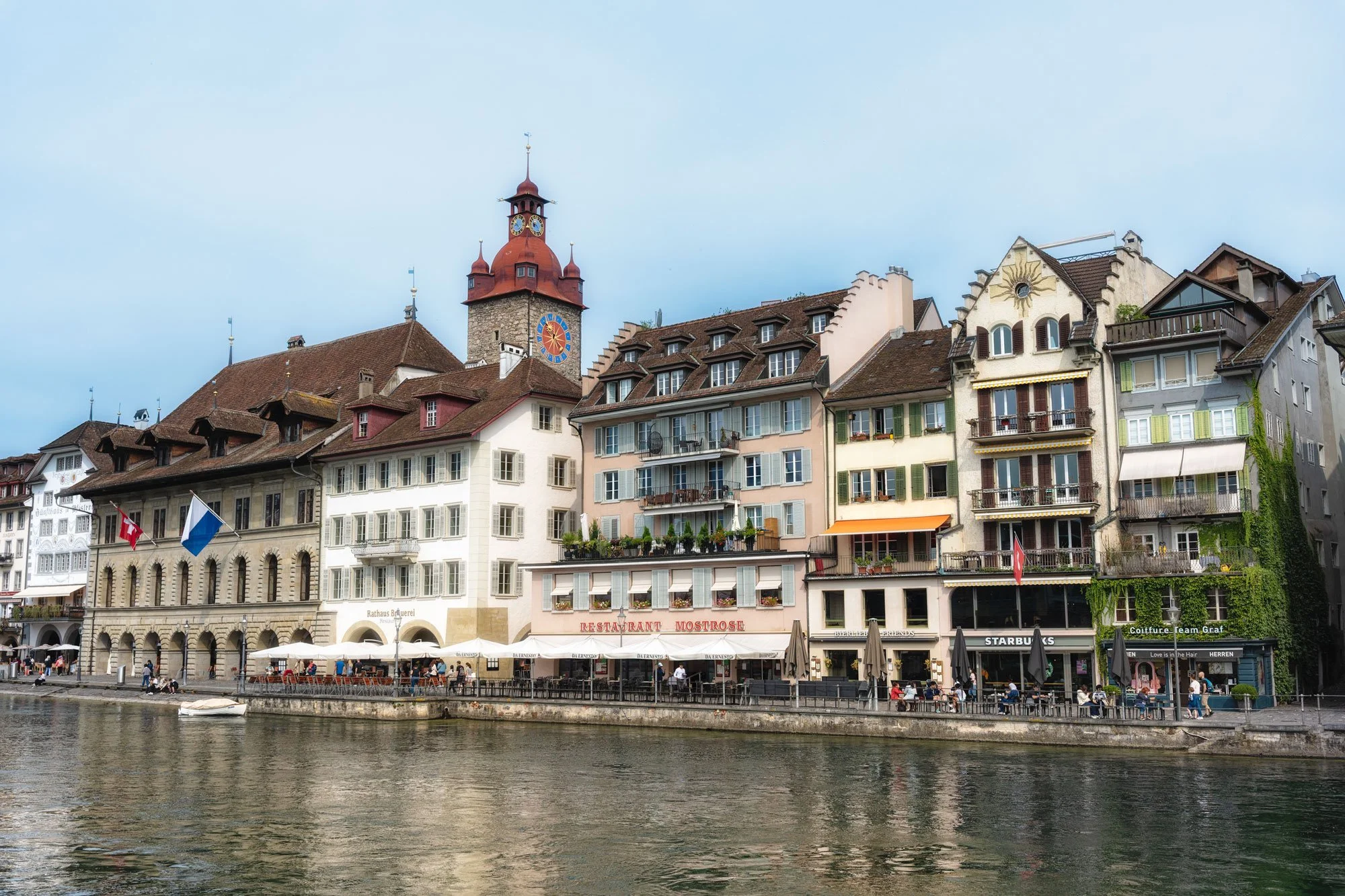 Photo of a historic European waterfront with colorful buildings, a church with a clock tower, and outdoor cafes along the riverbank.