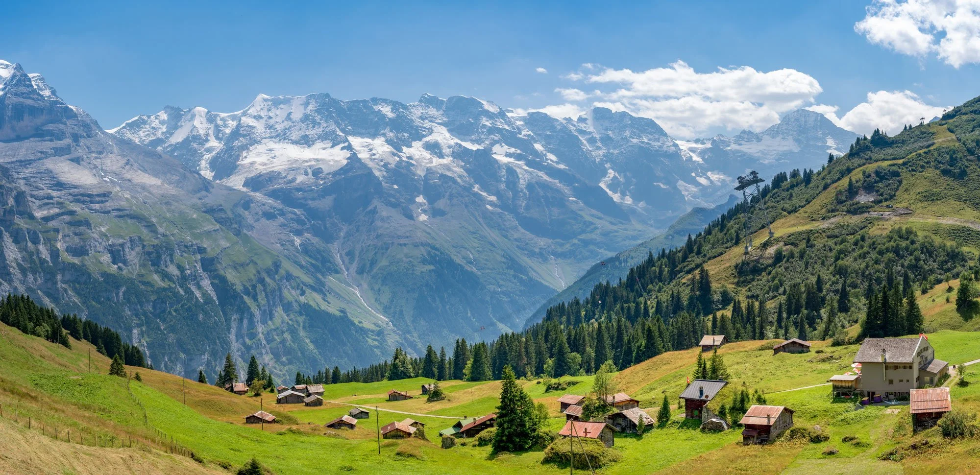 Scenic mountain village with houses on lush green hills, forested slopes, and snow-capped mountains under a partly cloudy blue sky.
