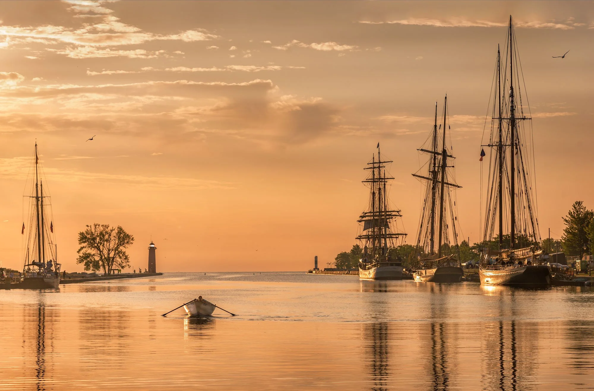 Sunset over a harbor with sailboats docked, a small boat on the water, trees, two lighthouses, and birds flying in the sky.