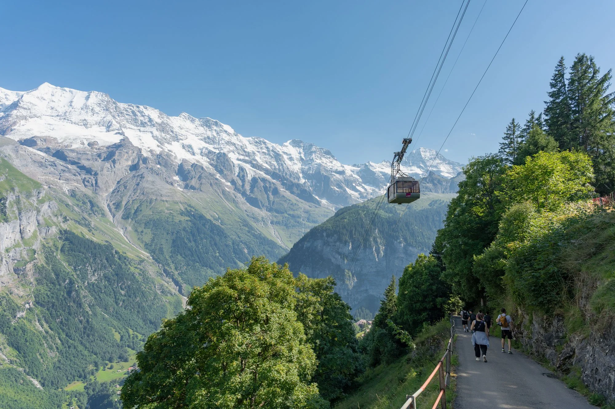 Tourists walking on a mountain pathway with a cable car overhead, snow-capped peaks in the background, and green trees lining the trail.
