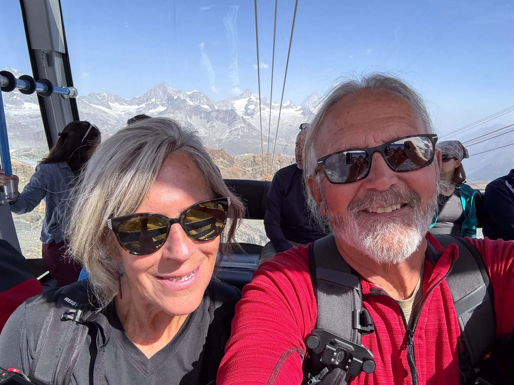 Smiling older couple taking a selfie on a cable car with snow-capped mountains in the background.