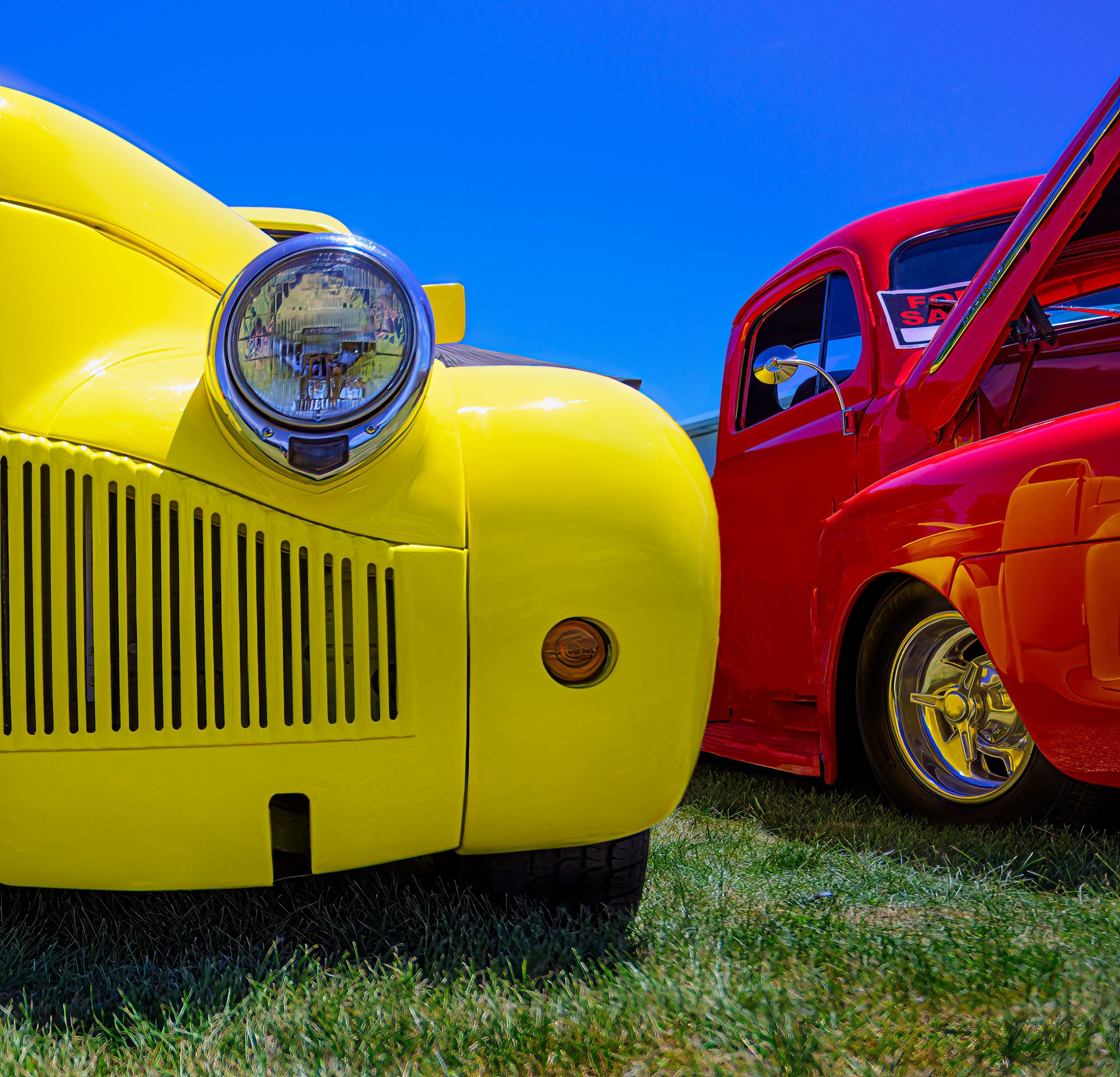 Close-up of two vintage cars parked on grass, one yellow and one red, with clear blue sky in the background.