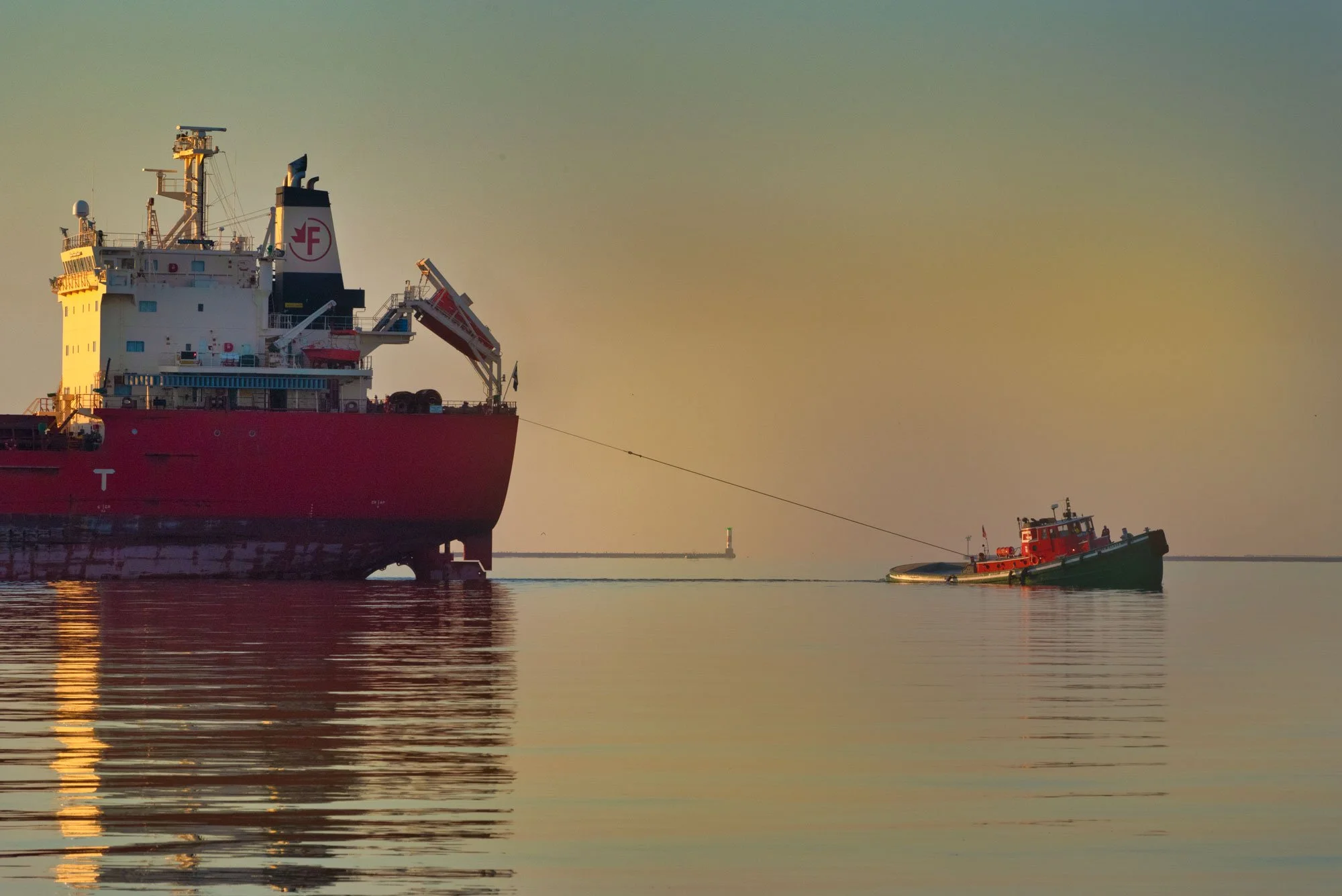 A large red and white ship with a smaller tugboat in front, towing it in calm water during sunset or sunrise.