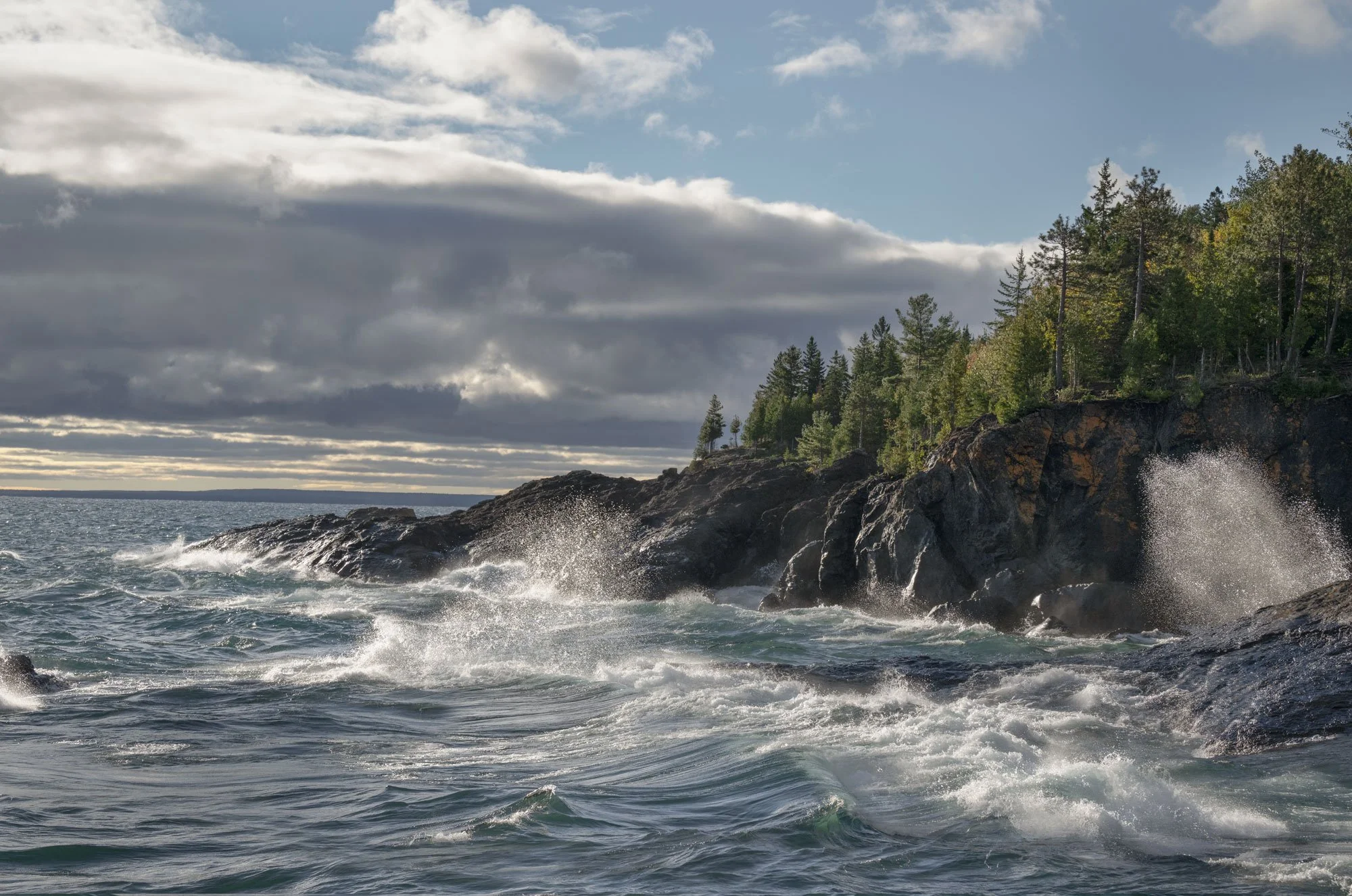 Waves crashing against rocky shoreline with green trees, cloudy sky