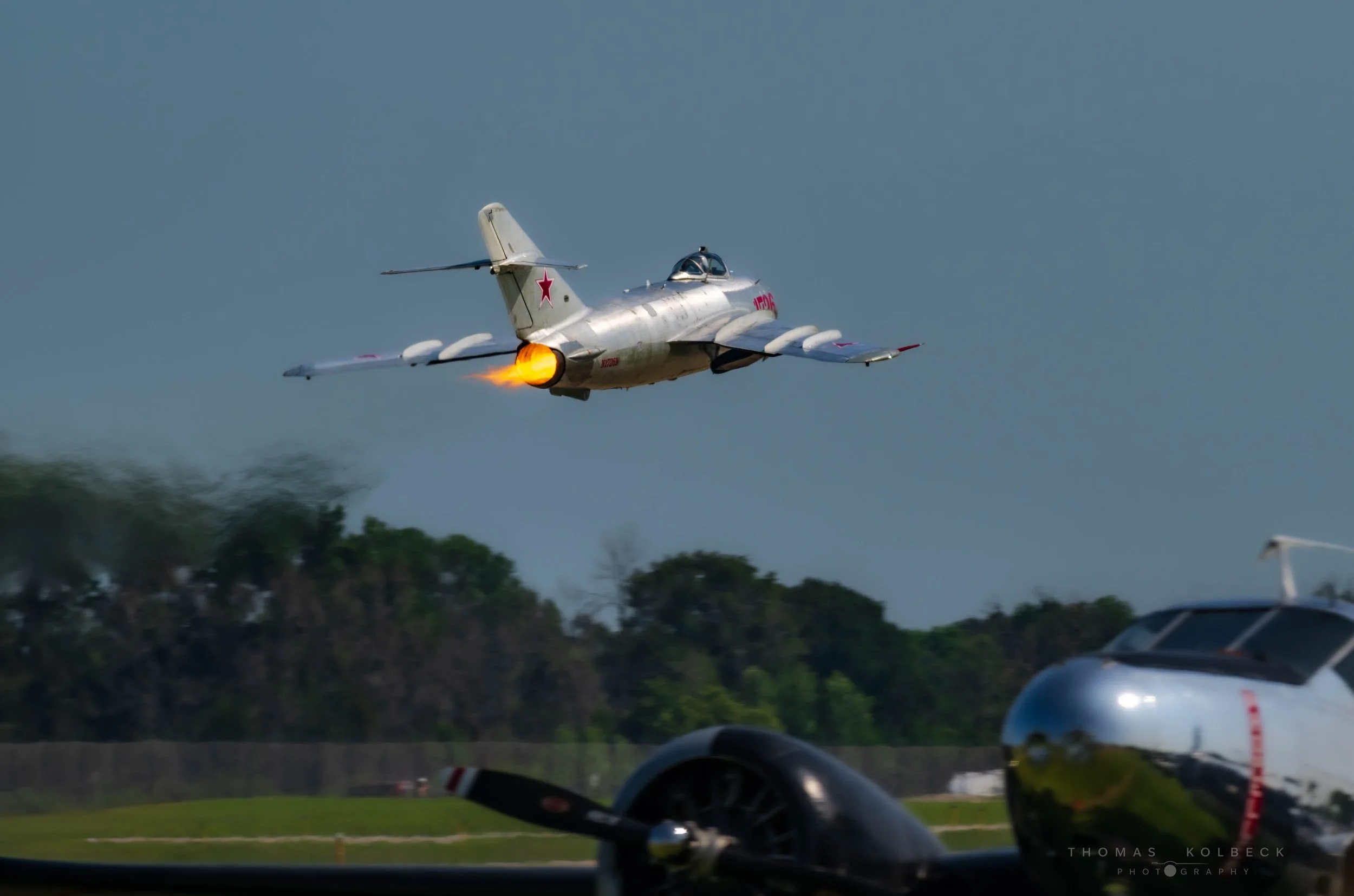A jet aircraft taking off from a runway with afterburner flames visible, and another aircraft visible in the foreground.