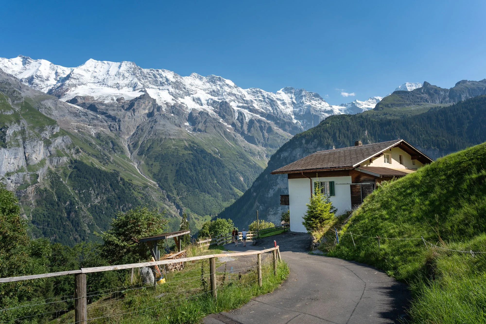 A mountain landscape with snow-capped peaks, green forested slopes, and a winding road leading to a house with a sloped roof, surrounded by grassy hills and a clear blue sky.