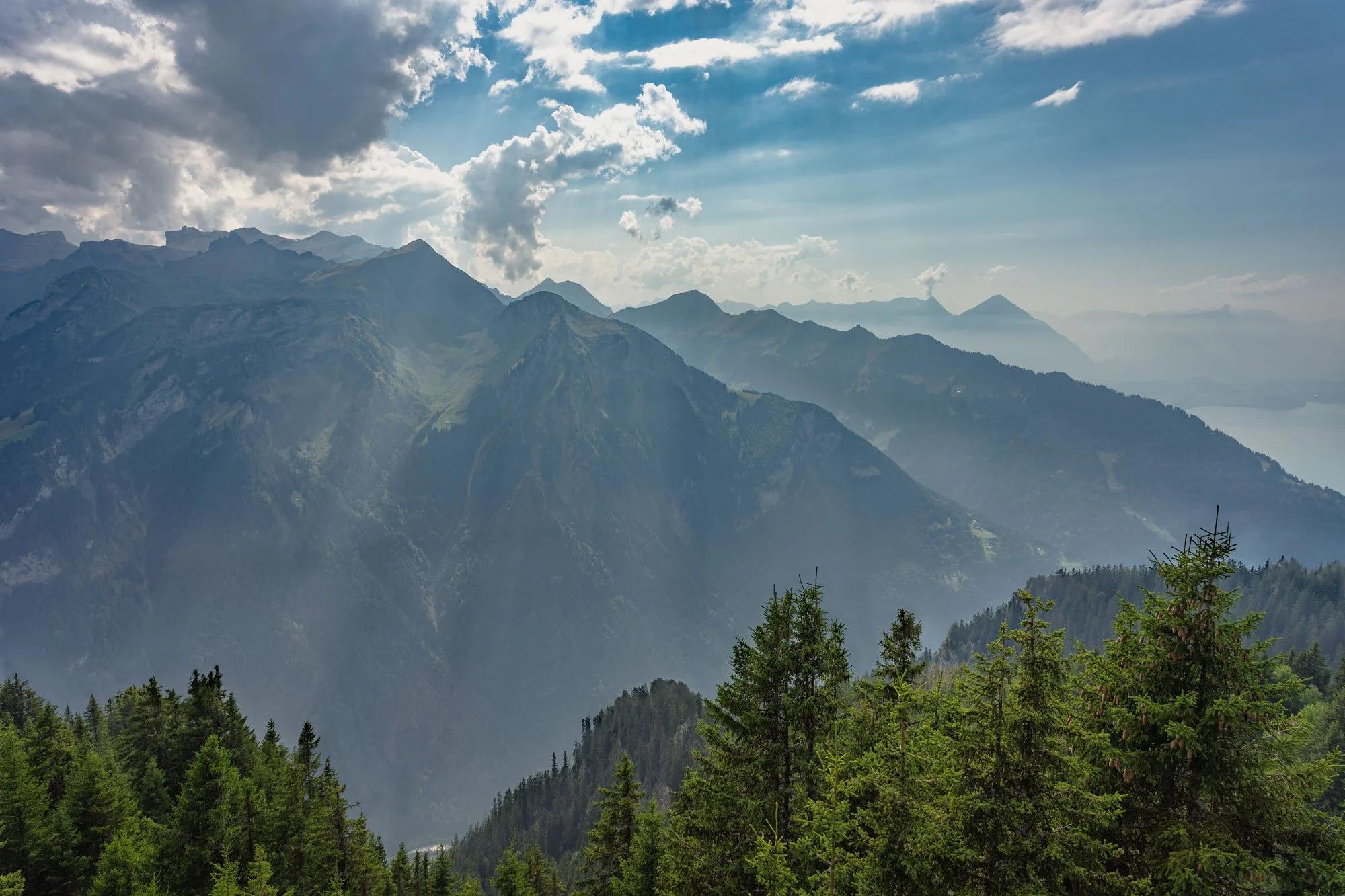 Mountain landscape with forested slopes, distant peaks, and a partly cloudy sky.