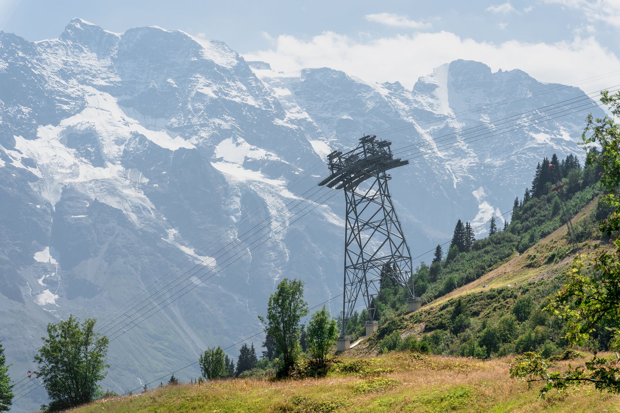 A mountain landscape with snow-capped peaks, green hills, trees, and a tall electrical transmission tower with power lines.