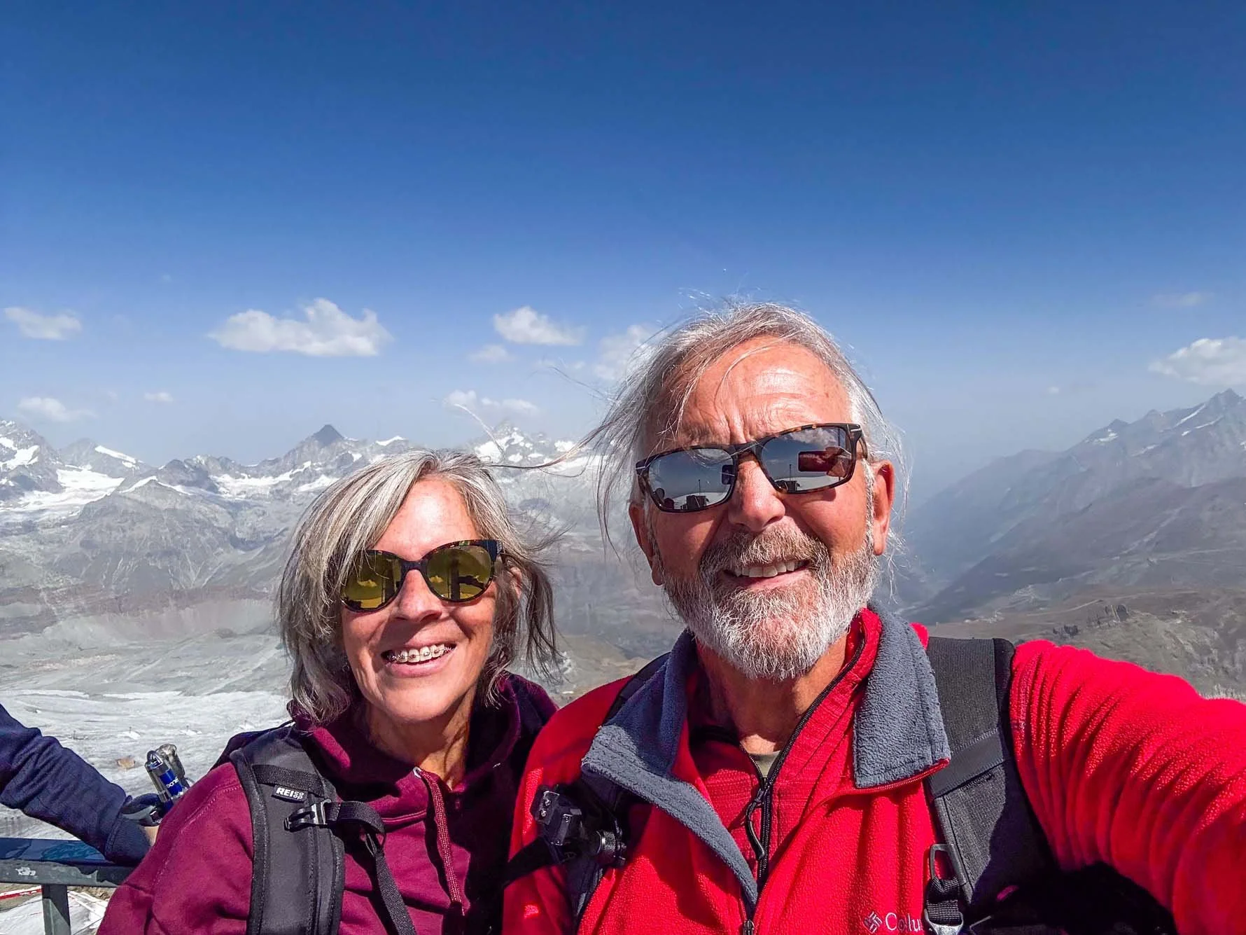 A smiling older man and woman wearing sunglasses in a mountainous landscape with snowy peaks and blue sky.