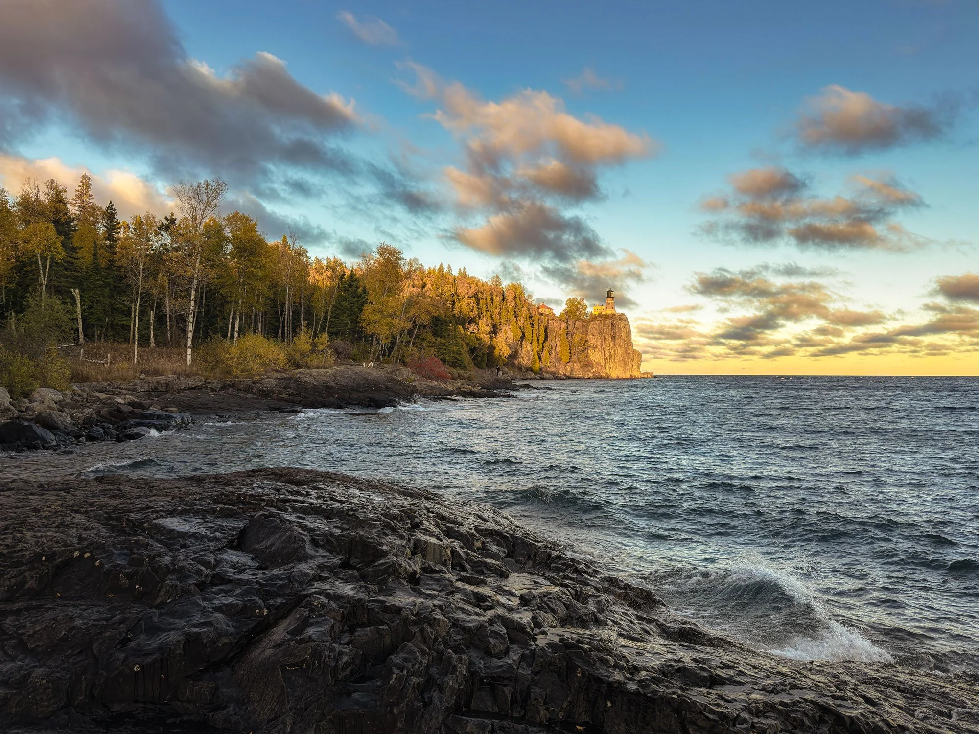 A rocky shoreline by the water with a dense forest and a lighthouse on a cliff in the background, under a partly cloudy sky during sunset.