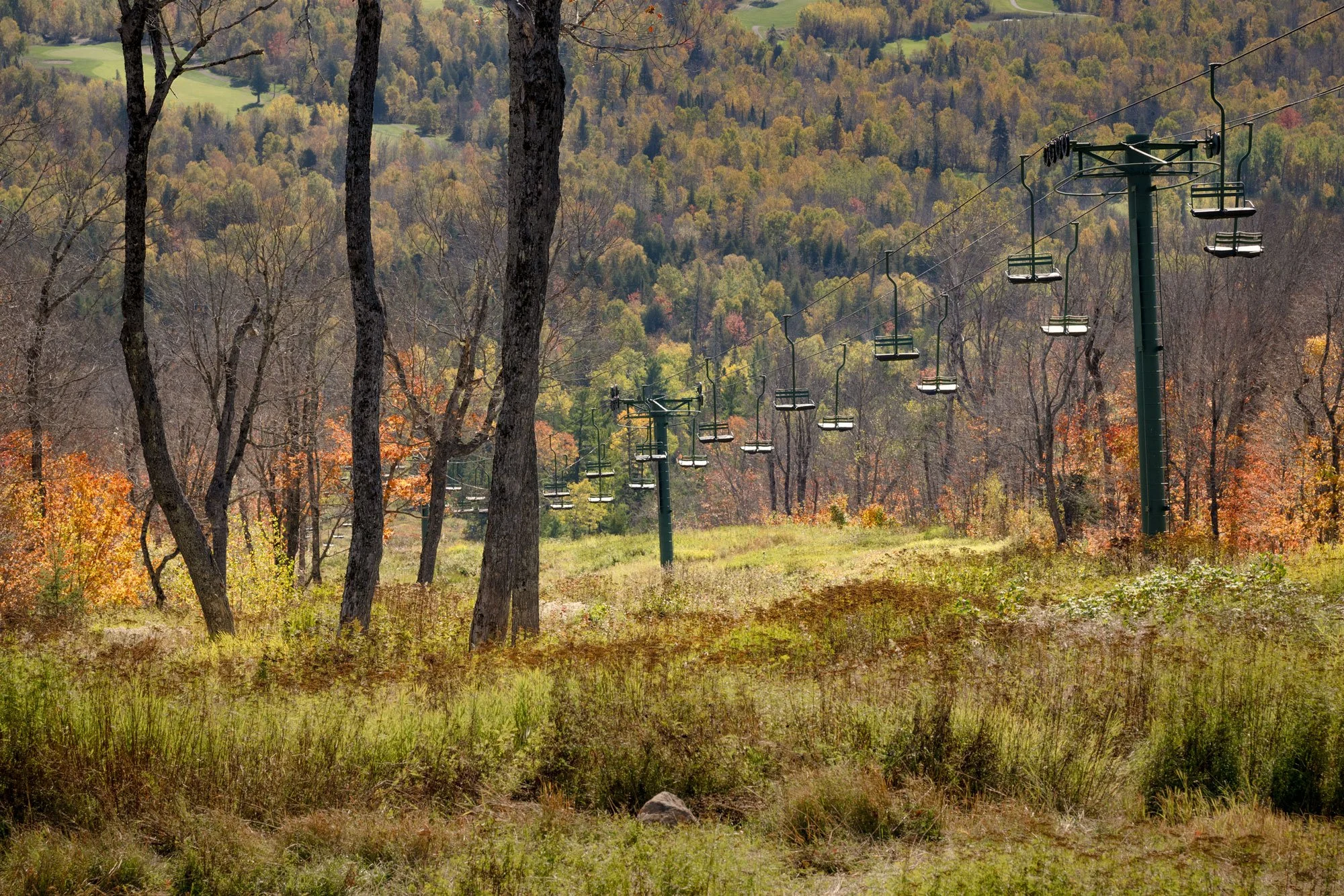 Empty ski lift chairs on a mountain trail surrounded by trees with autumn foliage.