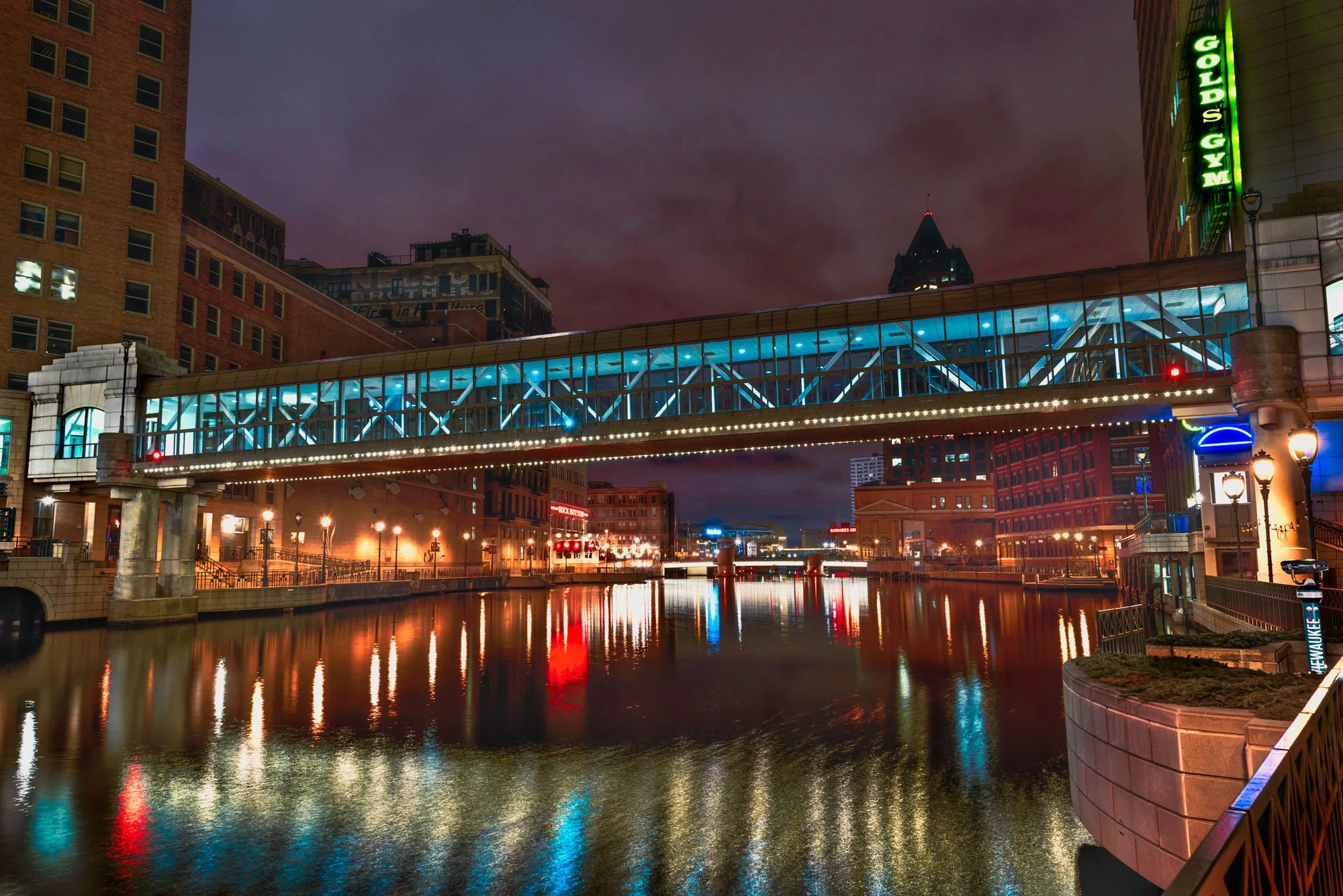 Cityscape at night with a bridge over a river, buildings with illuminated signs, and reflections in the water.