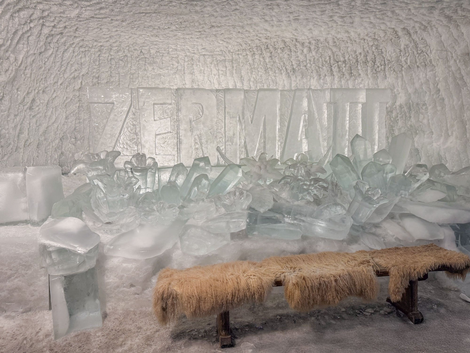 Ice sculpture display with the word 'ZEREMATI' carved in ice behind a pile of ice blocks, with a brown furry bench in front.