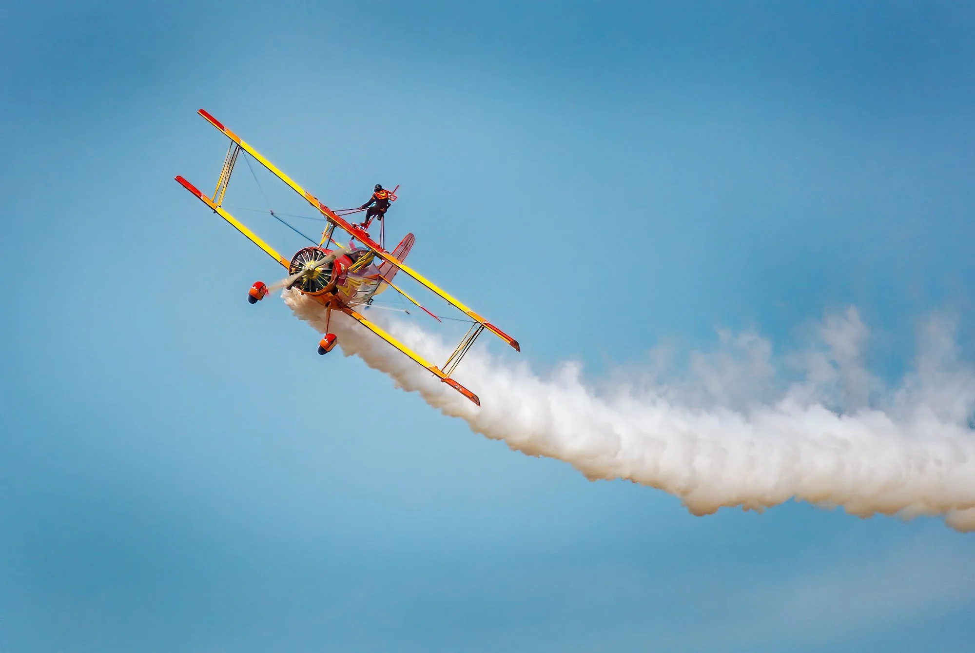 Aerial stunt pilot performing a loop in a bright yellow and red biplane, leaving white smoke trail against a blue sky.