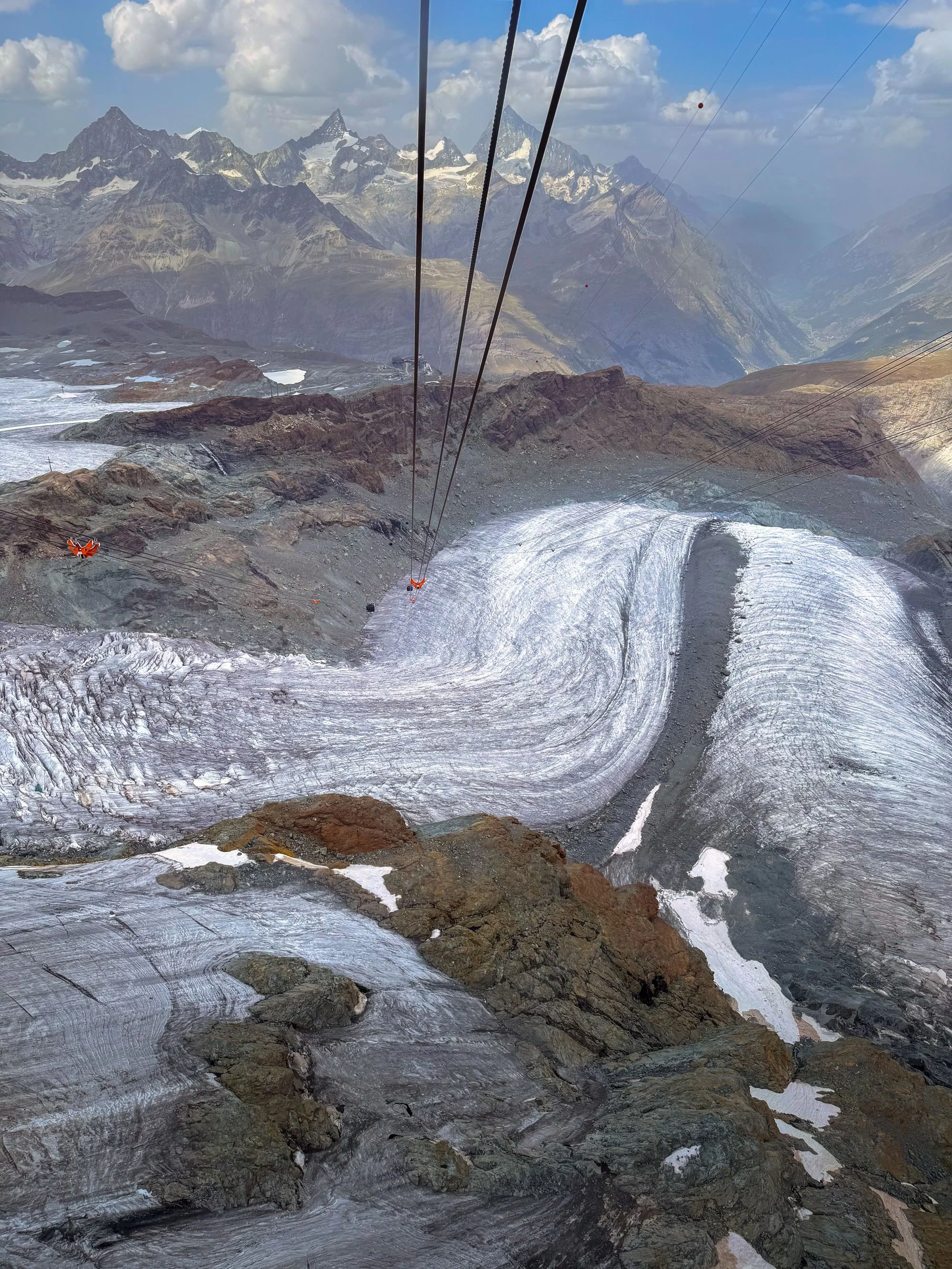 View from a mountain cable car showing snow and ice on rocky terrain, with distant snow-capped mountains under a partly cloudy sky.