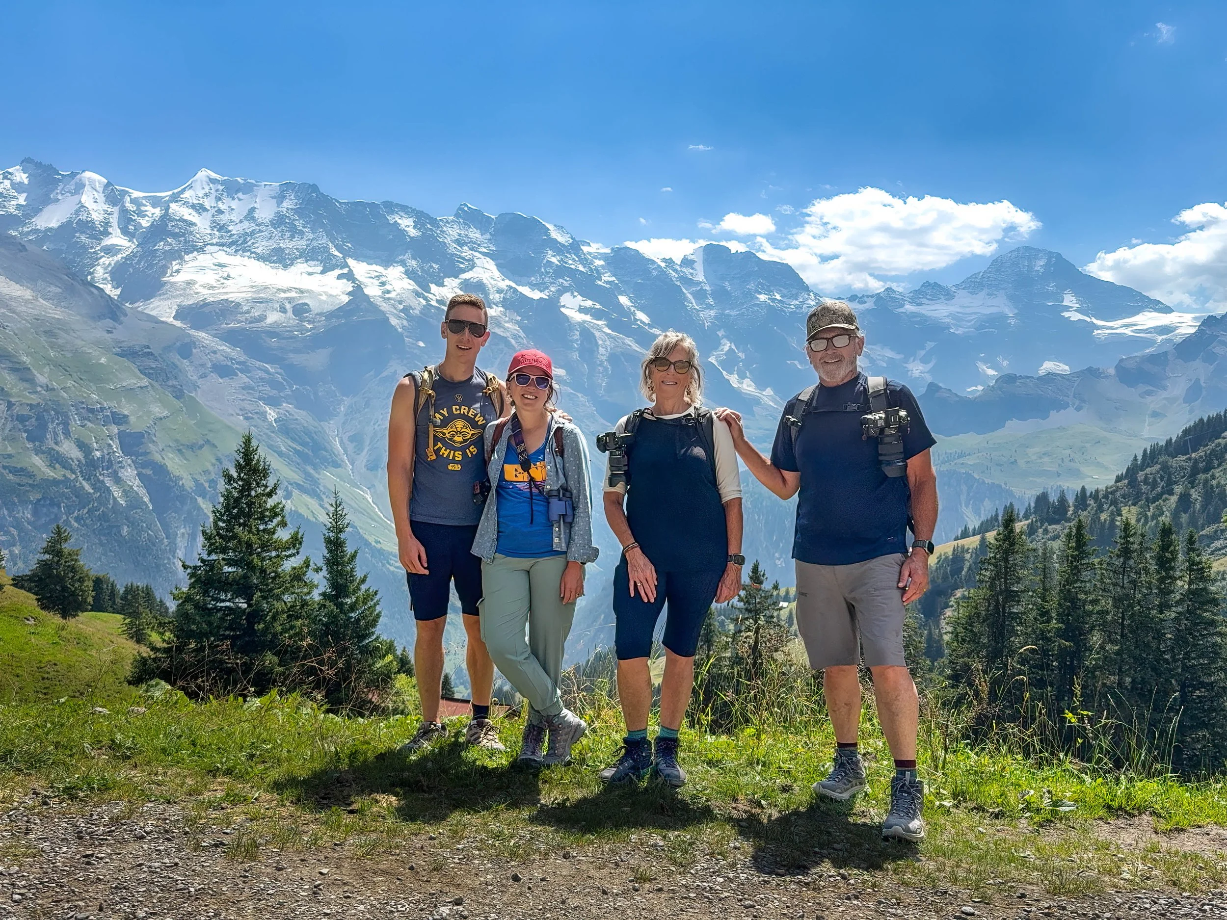 Four people standing on a grassy trail with snow-capped mountains and trees in the background, enjoying a sunny day in a scenic outdoor setting.