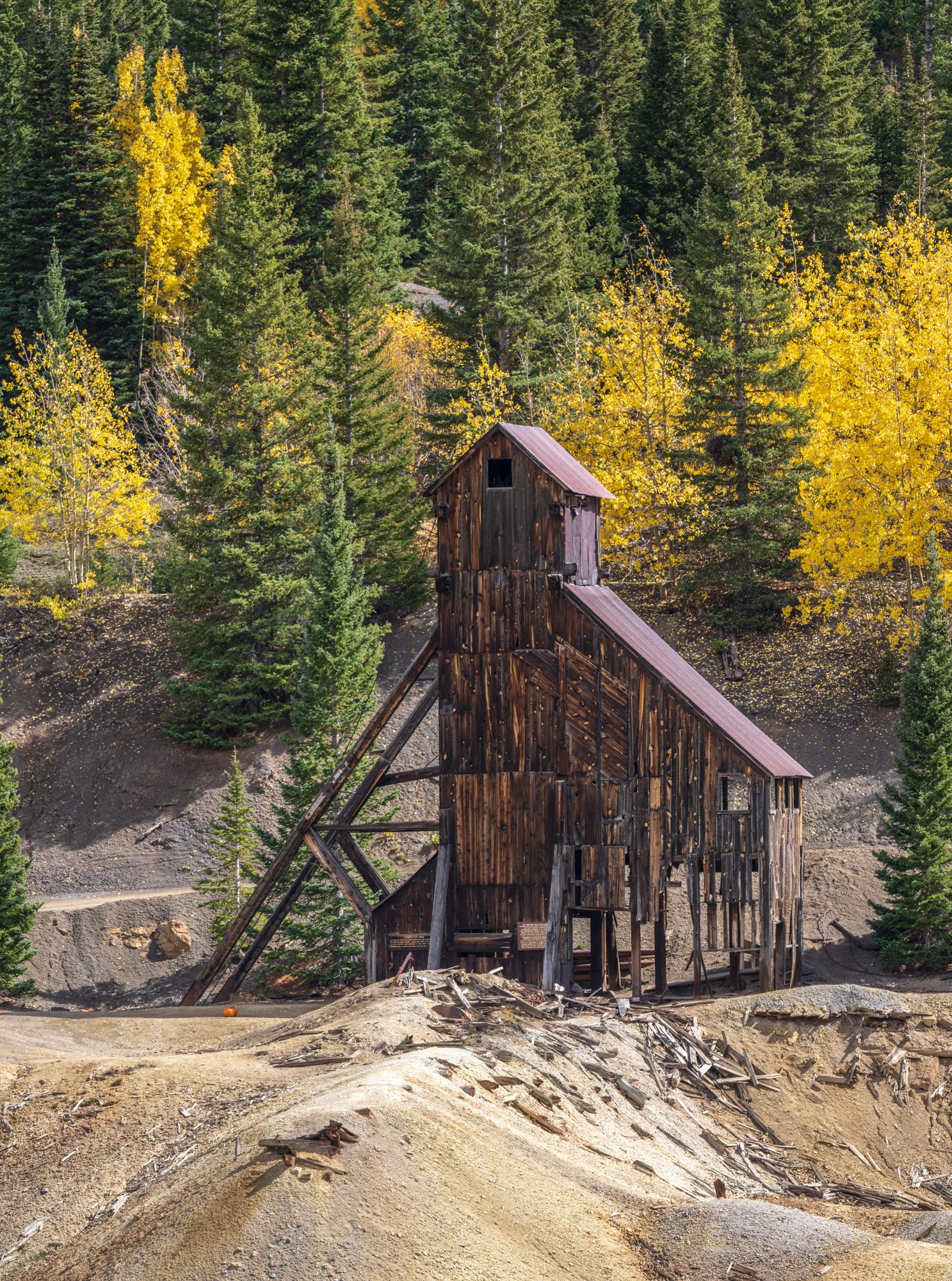 An old wooden mine shaft building surrounded by trees with yellow and green autumn foliage, in a mountainous area.