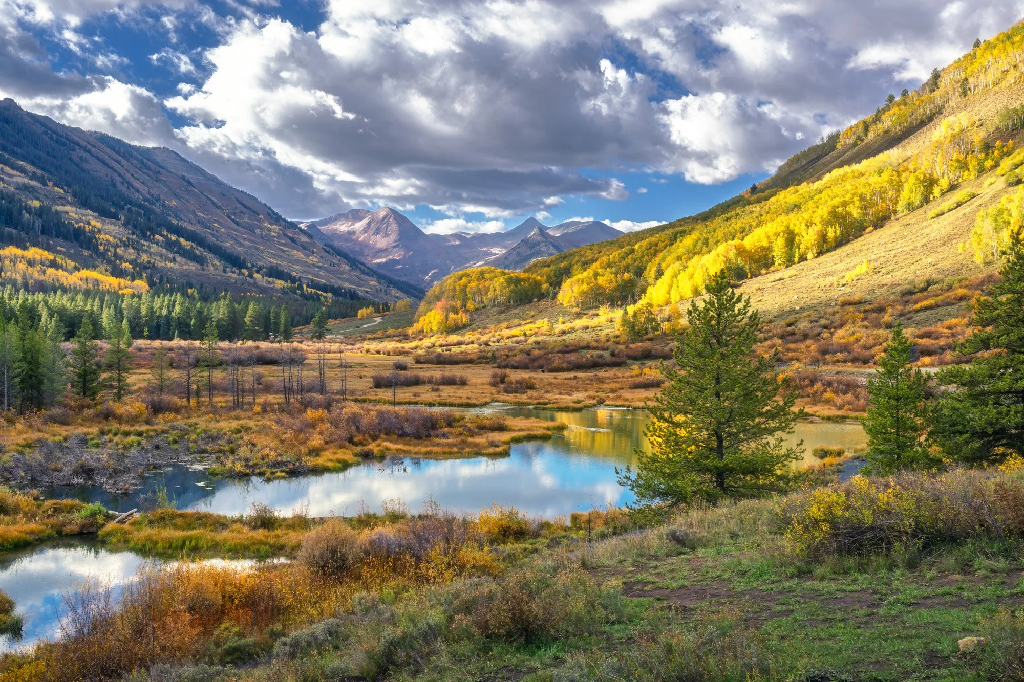 A scenic mountain landscape with colorful autumn foliage, a river flowing through the valley, and cloud-covered mountains in the background.