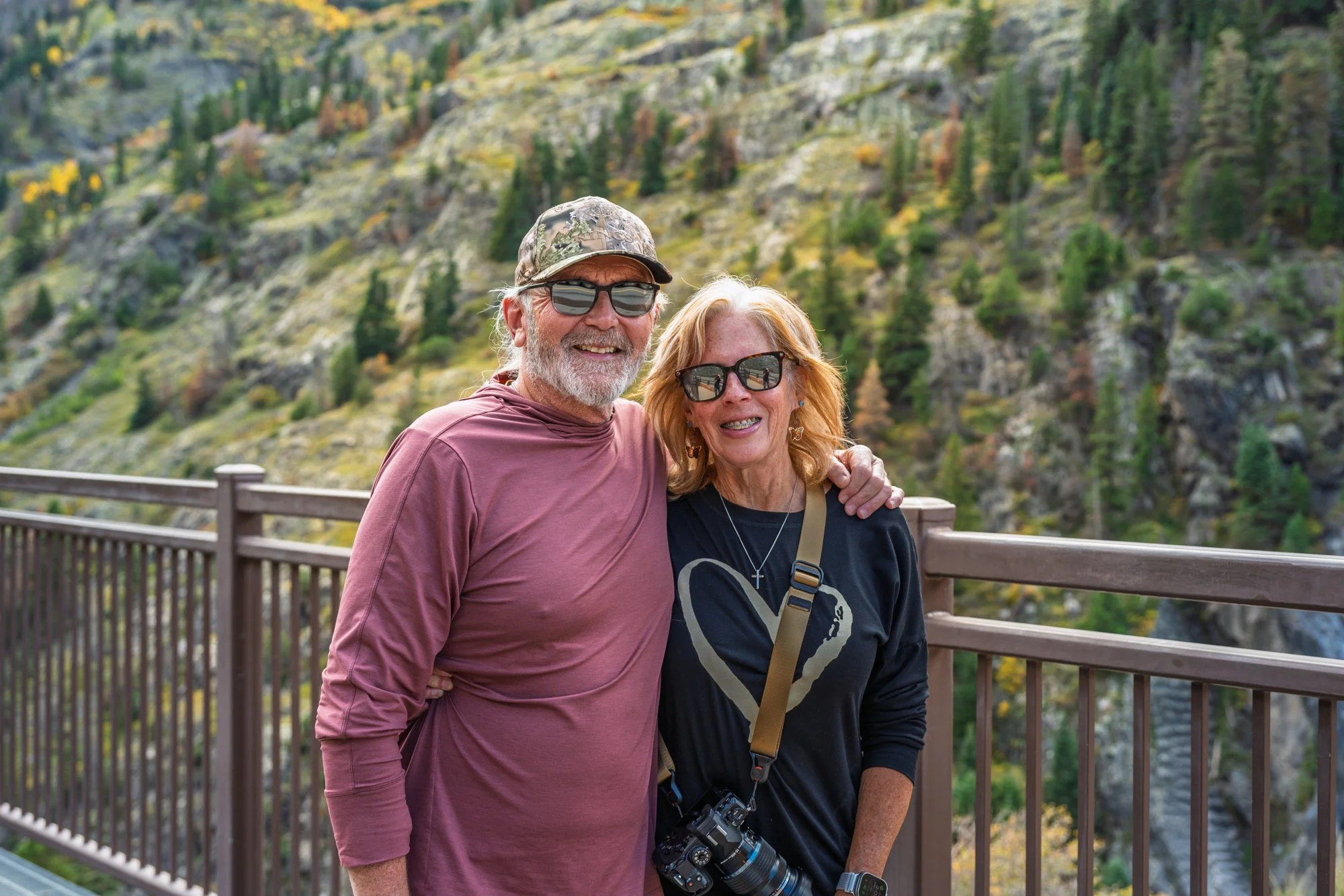 An elderly man and woman standing together outdoors on a wooden railing bridge with a mountainous landscape covered in trees in the background. Both are wearing sunglasses and smiling, the man has white hair, a beard, a camouflage cap, and a reddish 
