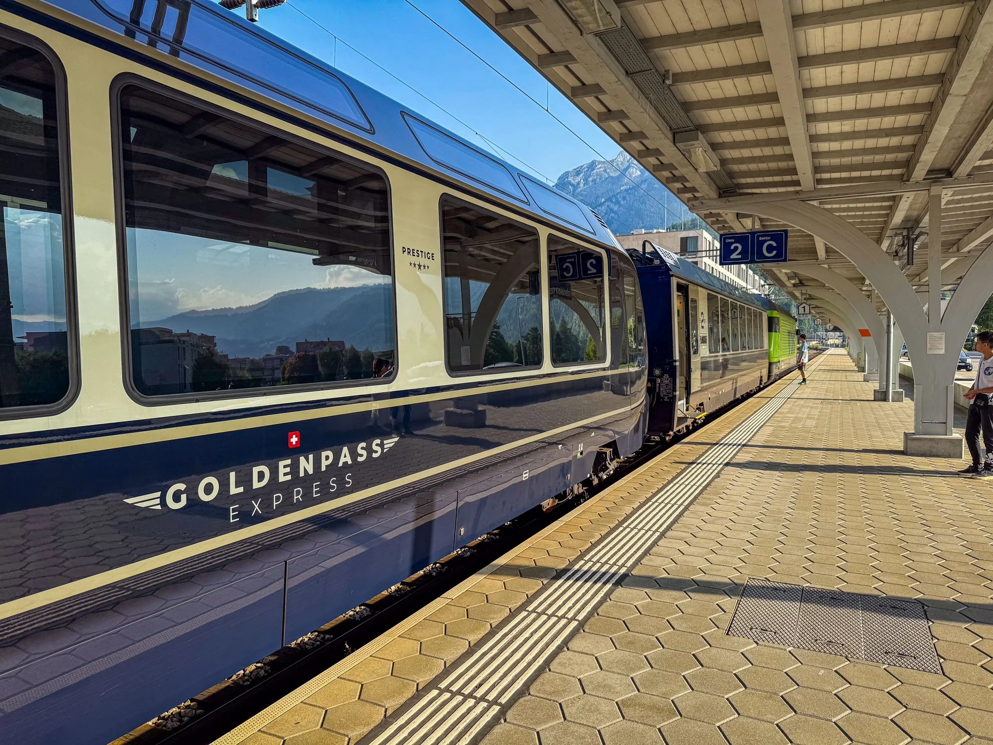 A Blue and cream train labeled 'GOLDENPASS EXPRESS' with mountainous scenery reflected in its window, at a station platform with a cover and signage indicating tracks 2 C, with a person nearby.