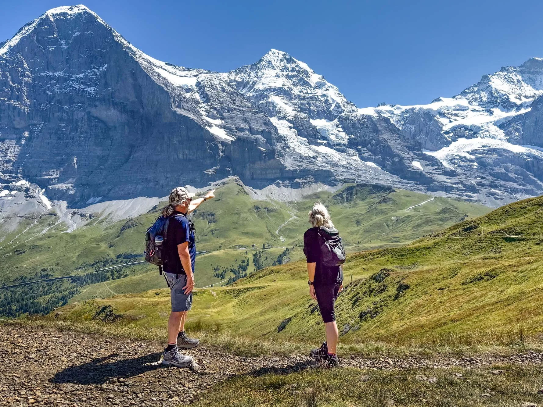 Two hikers standing on a trail in a green valley, with snow-capped mountains in the background, one pointing at the mountains.