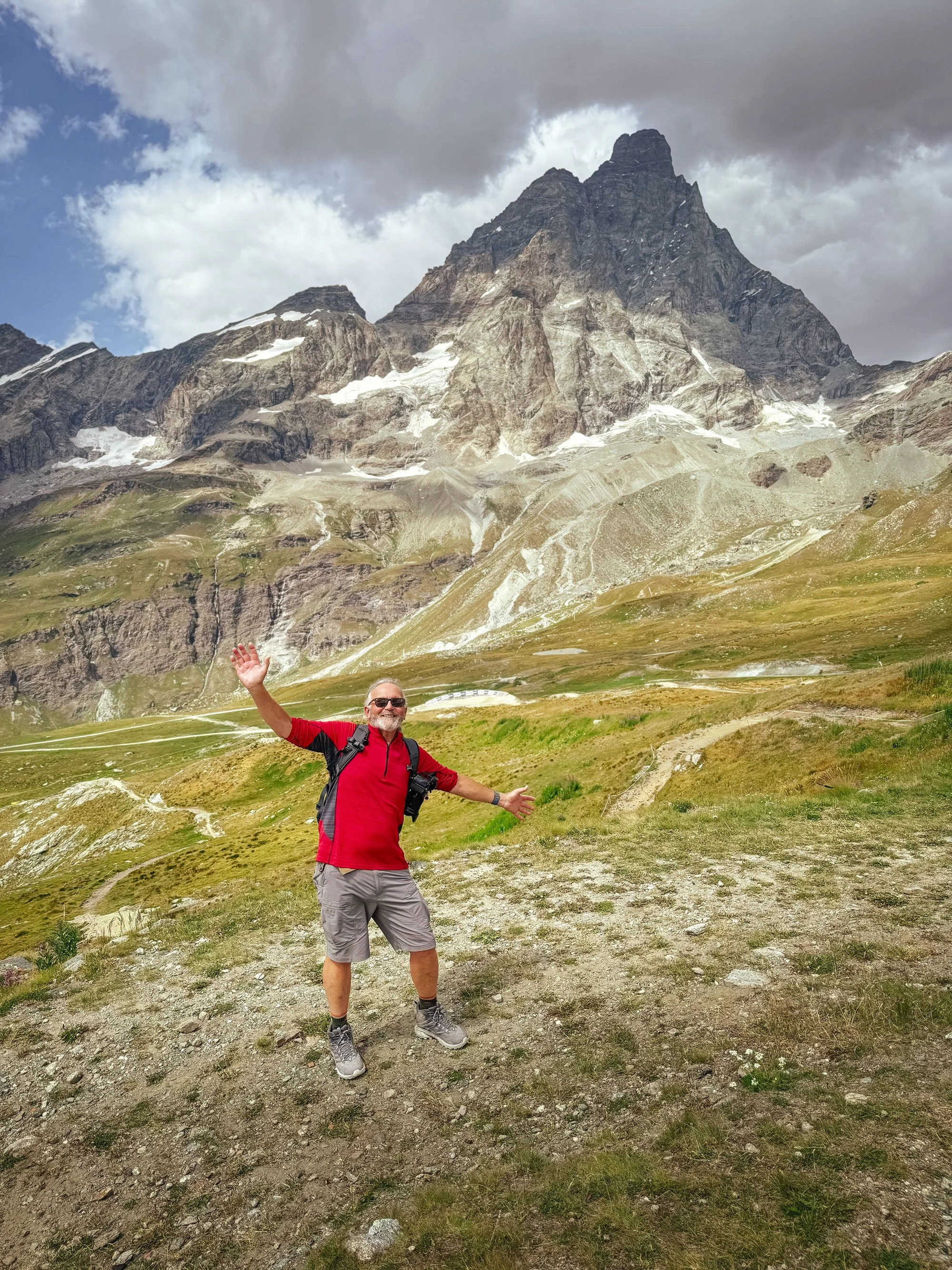 A man in a red shirt and gray shorts standing on a trail in a mountainous area in Switzerland, smiling and raising his arms, with tall mountain peaks and a partly cloudy sky in the background.