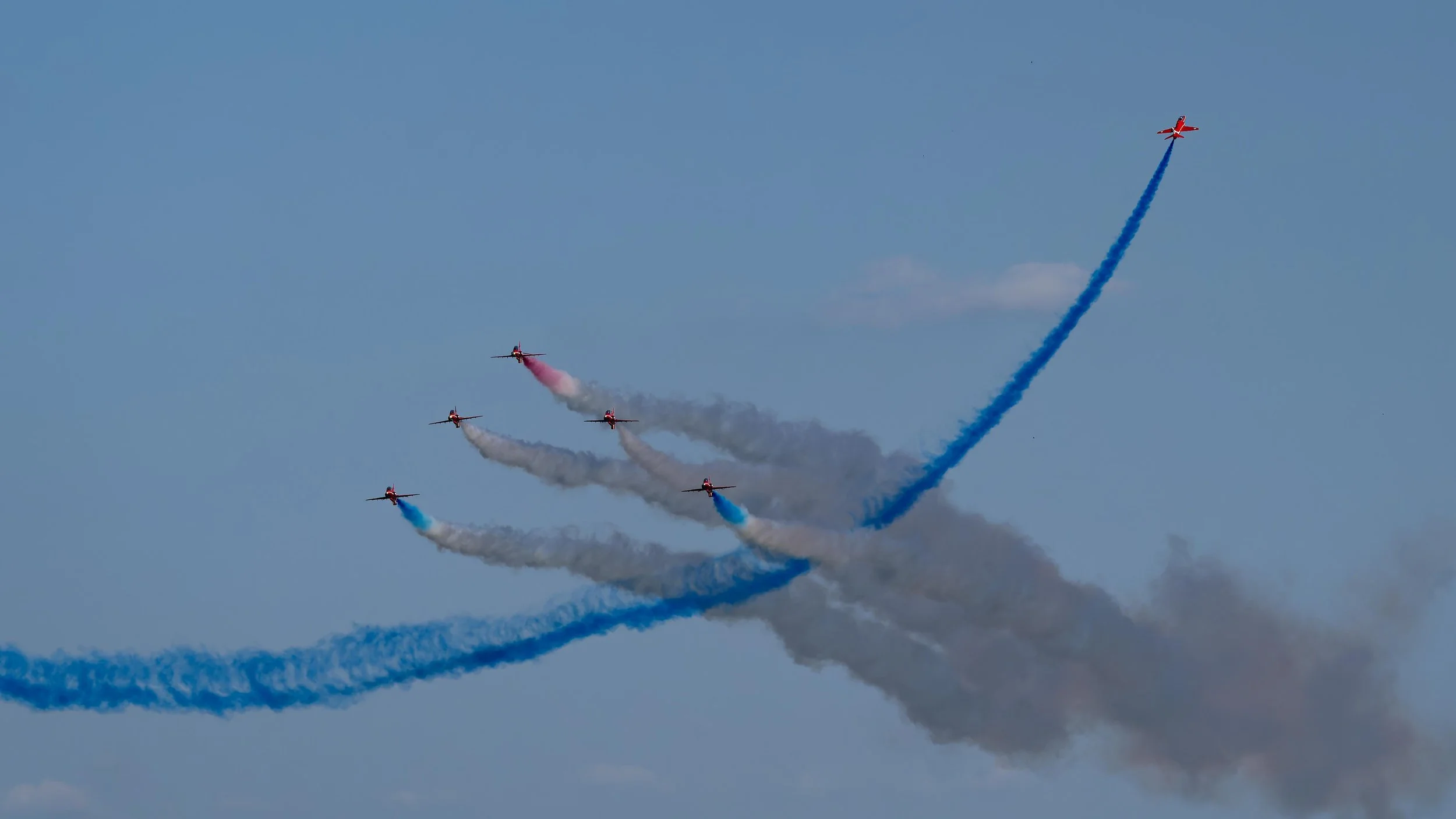 Six red aircrafts performing an aerial stunt, leaving colored smoke trails against a blue sky with some clouds.