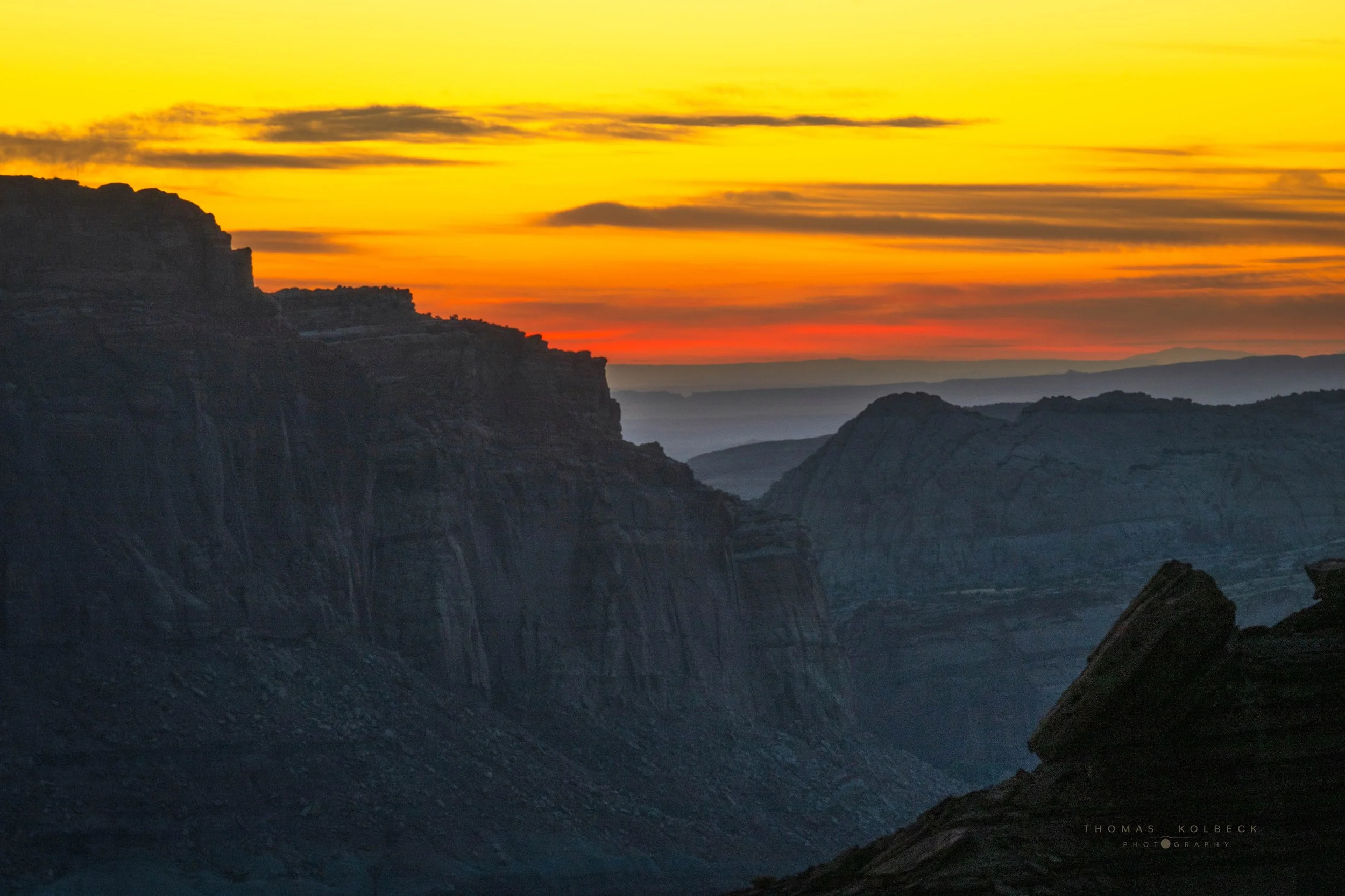 A mountain landscape during sunset with orange, yellow, and pink sky, dark rocky cliffs, and ridges in the distance.