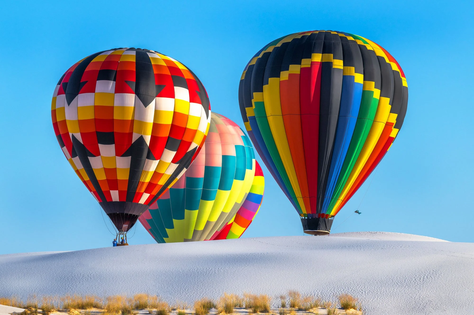Three colorful hot air balloons flying over a white sandy landscape with sparse grass, set against a clear blue sky.