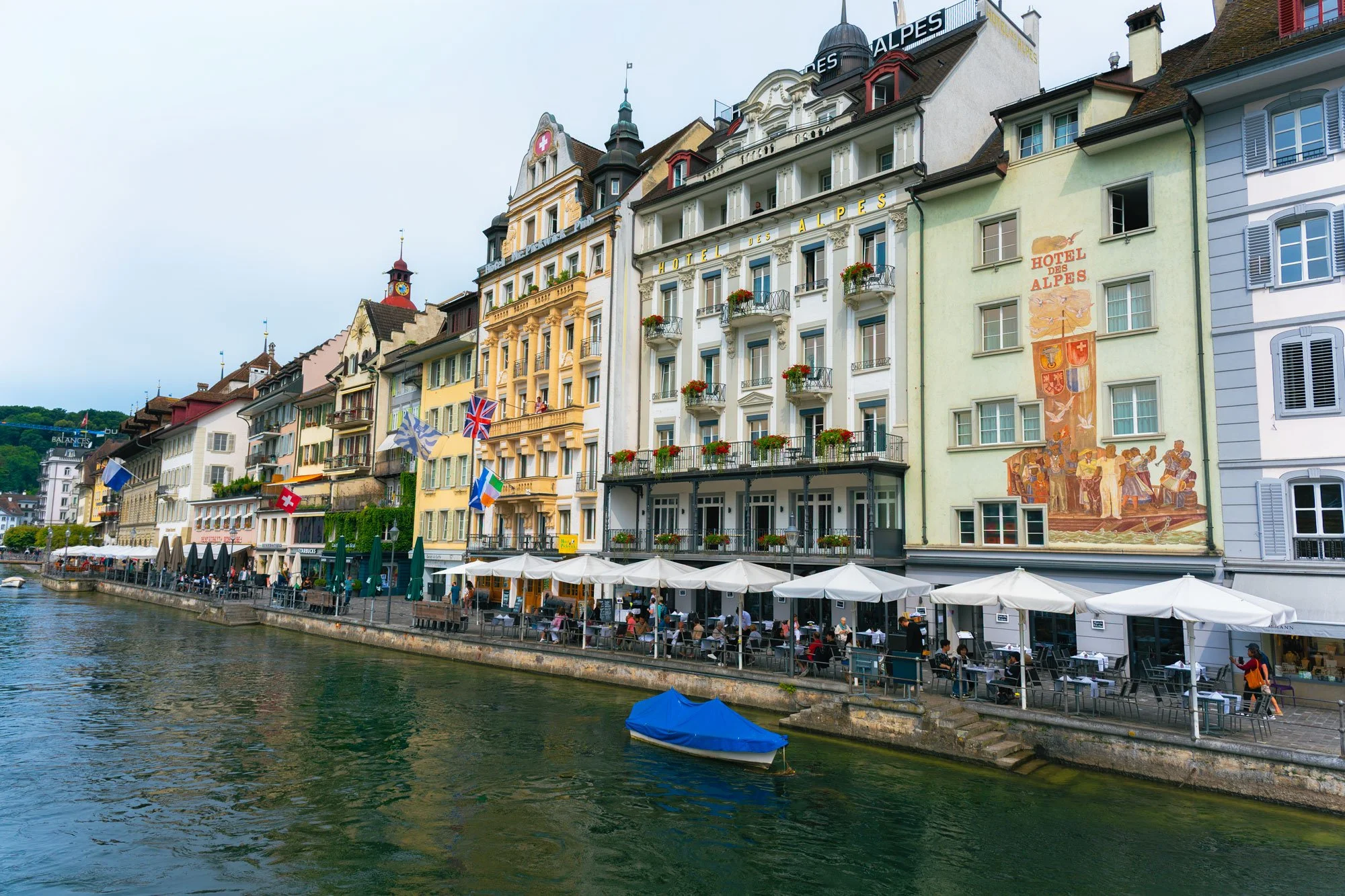 Colorful buildings with flags along a river with outdoor seating under white umbrellas and a small boat in the water.